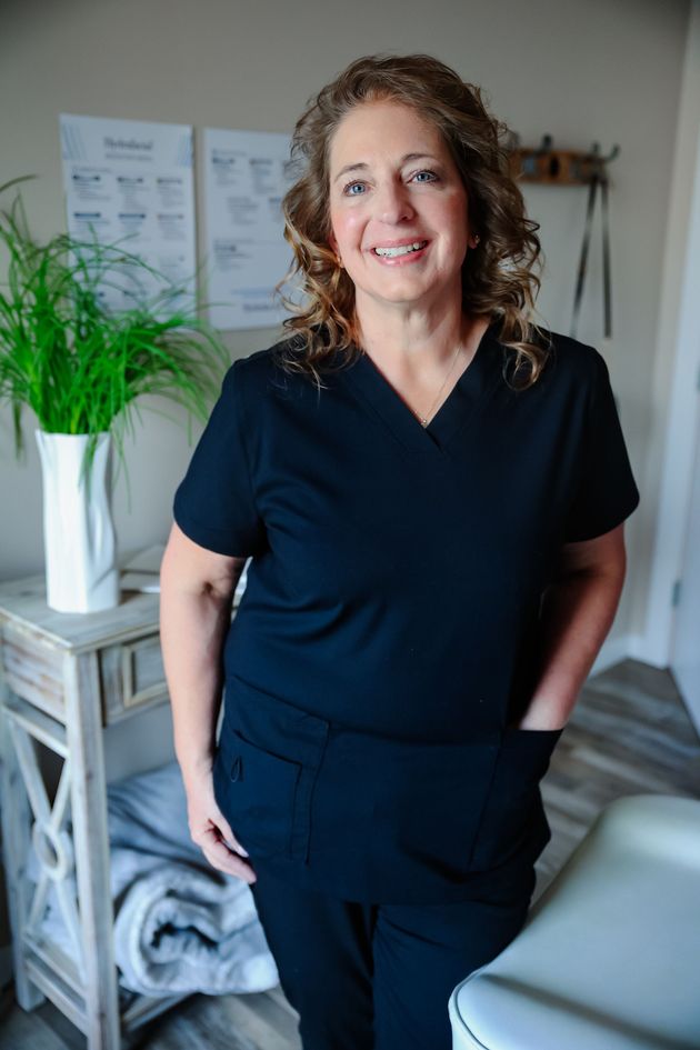 Woman standing in a modern room, wearing dark scrubs and smiling beside a leafy plant.