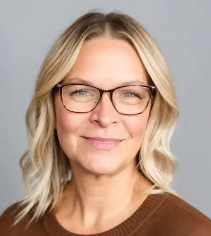 Smiling blonde person wearing glasses and a brown sweater against a gray background