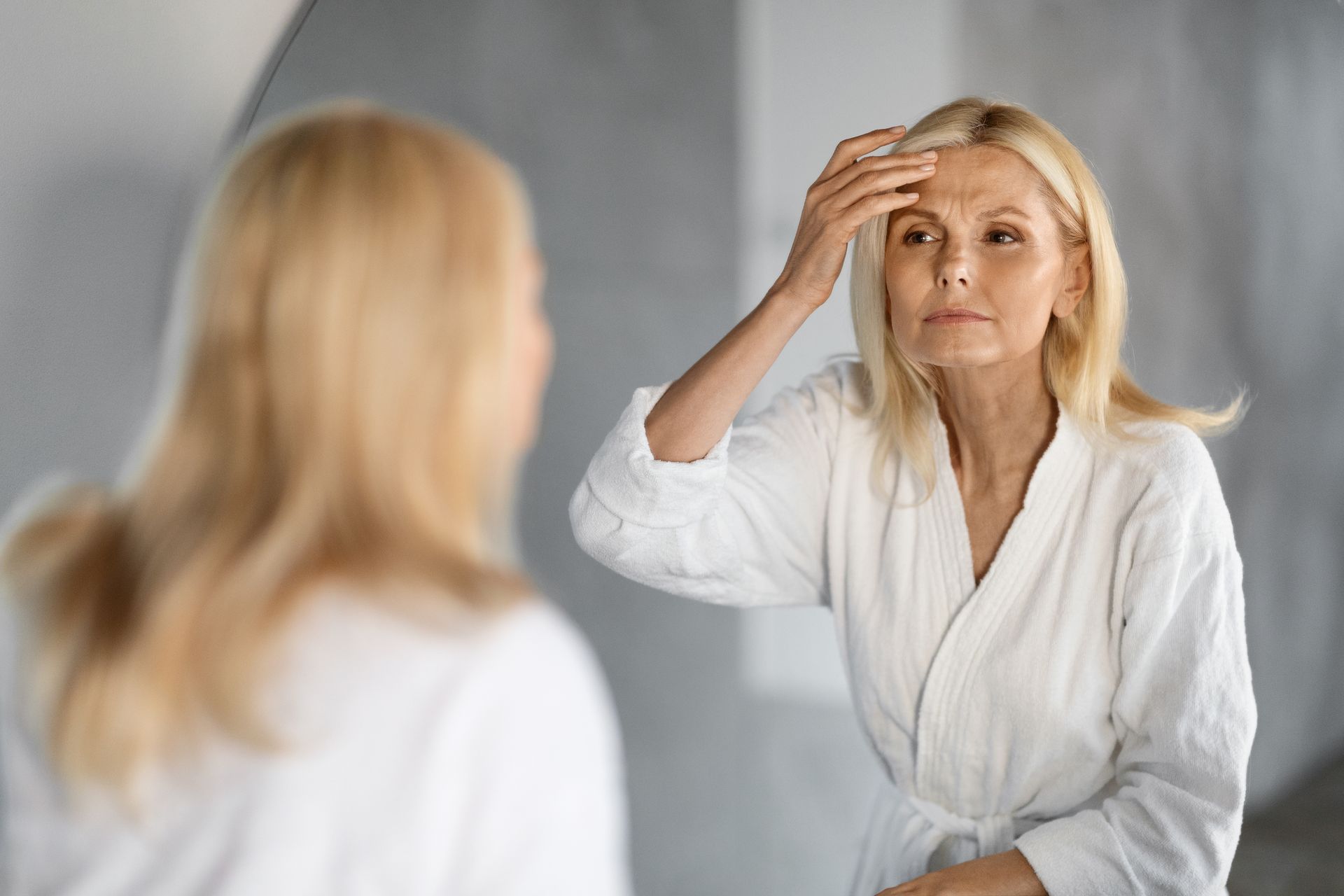 Woman in white robe looking in mirror, touching forehead, inspecting face.