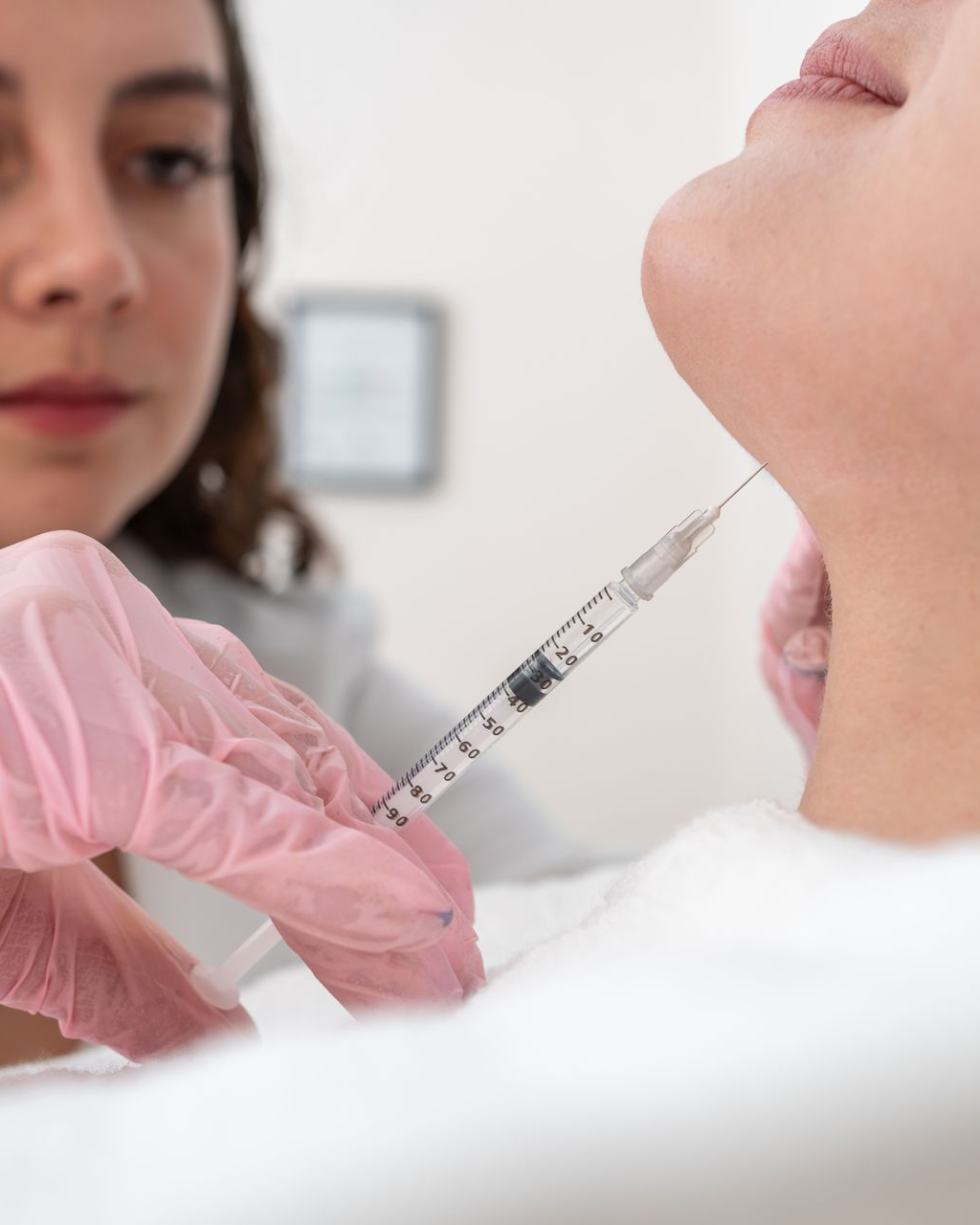 Hands in blue gloves injecting a syringe into a person's abdomen, on a white surface.