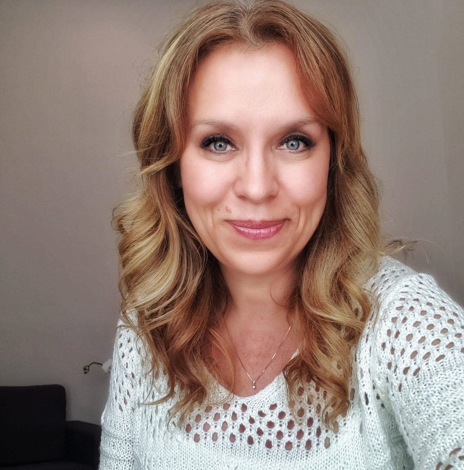 Smiling woman with wavy blonde hair wearing a white knit top against a neutral background