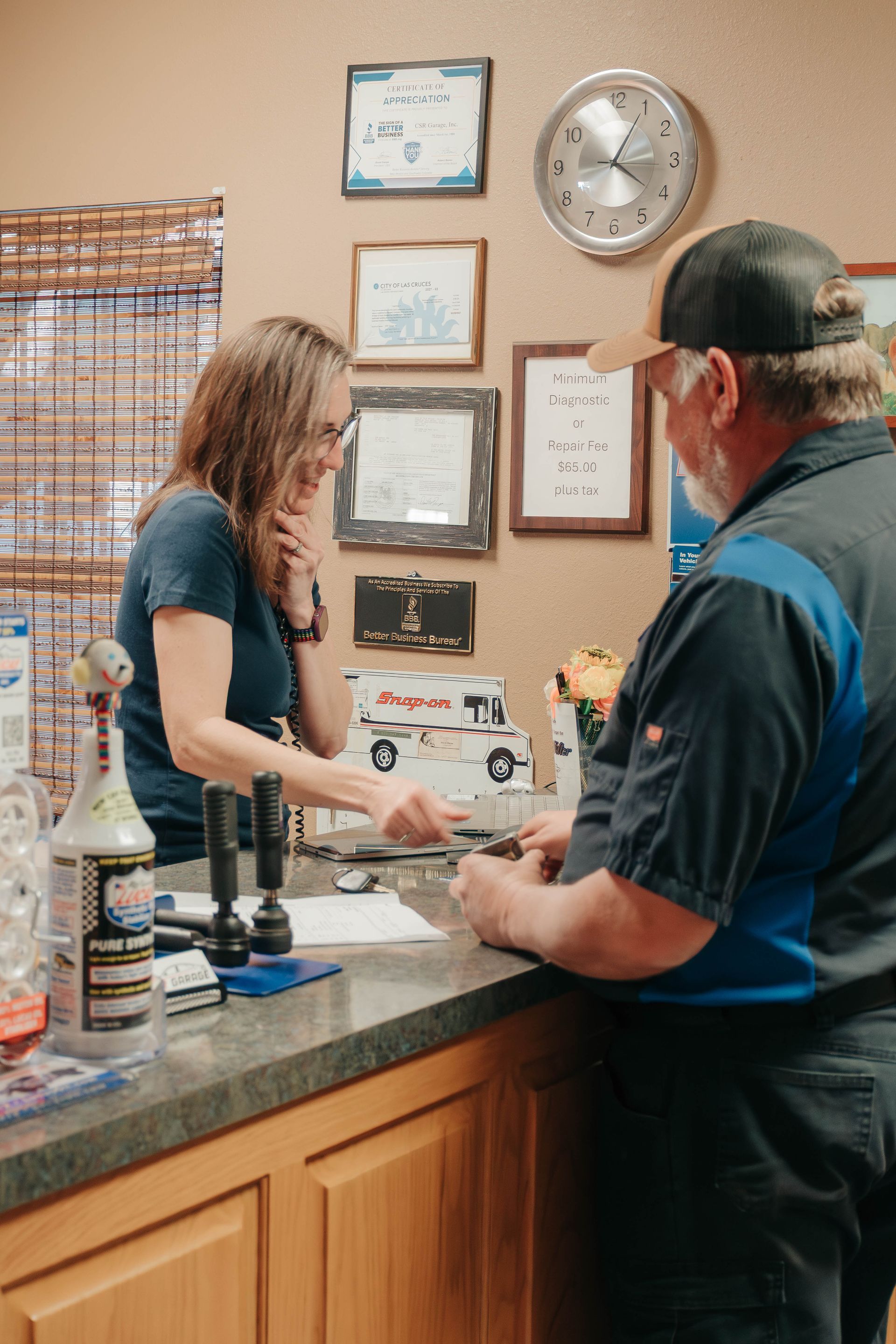 Two people at a counter in a cozy office, one writing while the other waits, with framed notes and a clock on the wall