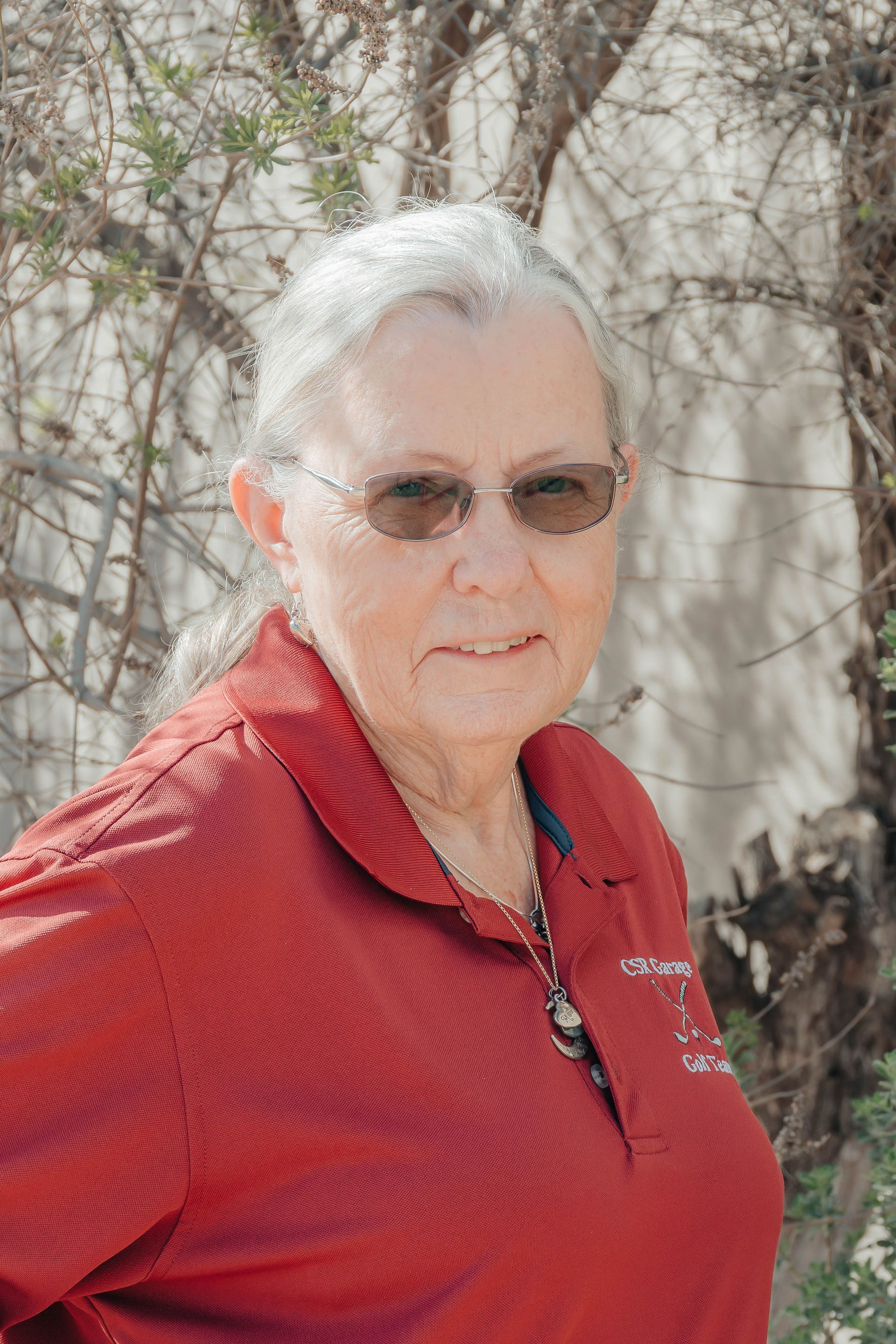 Person in a red polo shirt and sunglasses standing outdoors near a tree and wall.