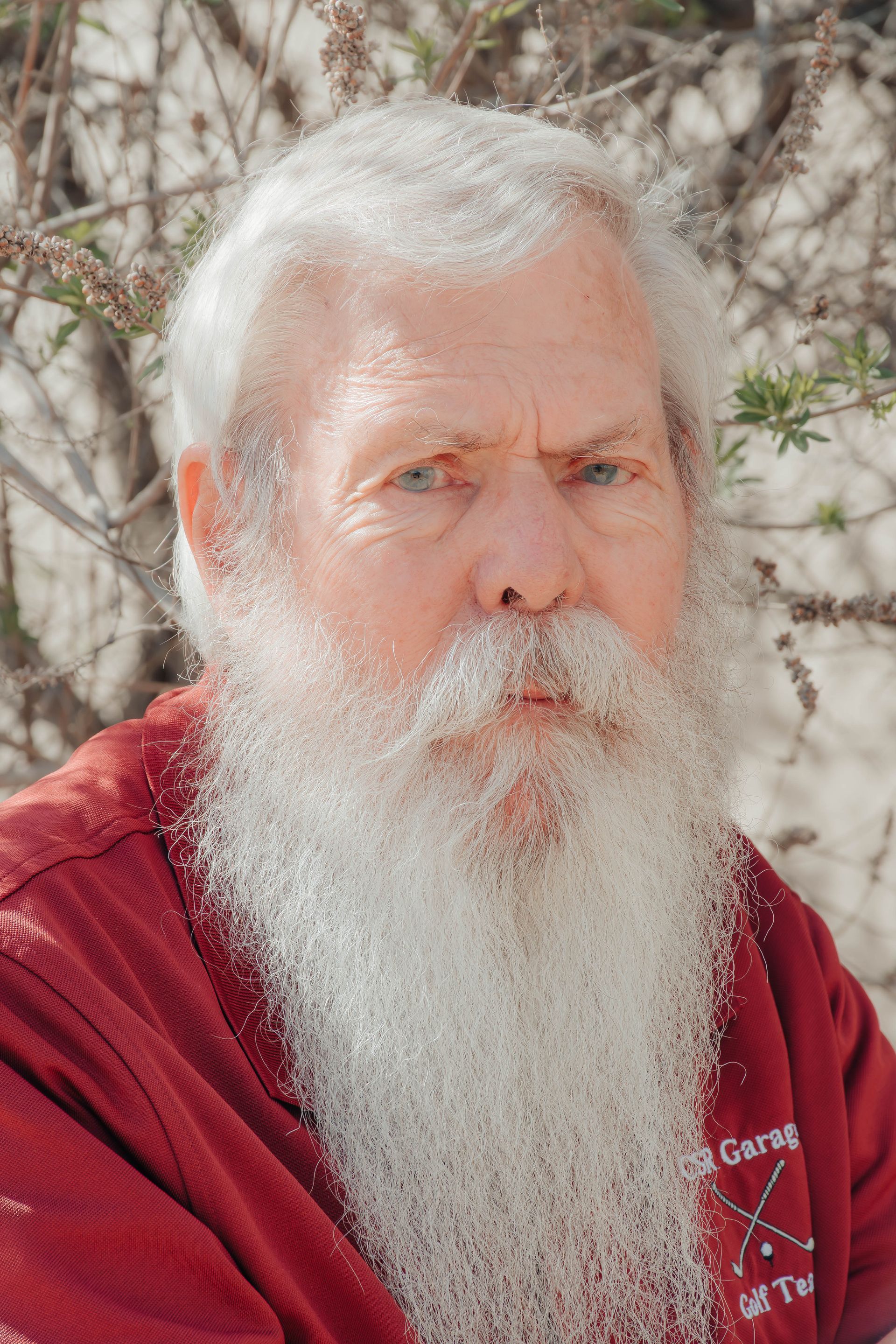 Portrait of a white-bearded man in a red shirt outdoors, with trees in the background.
