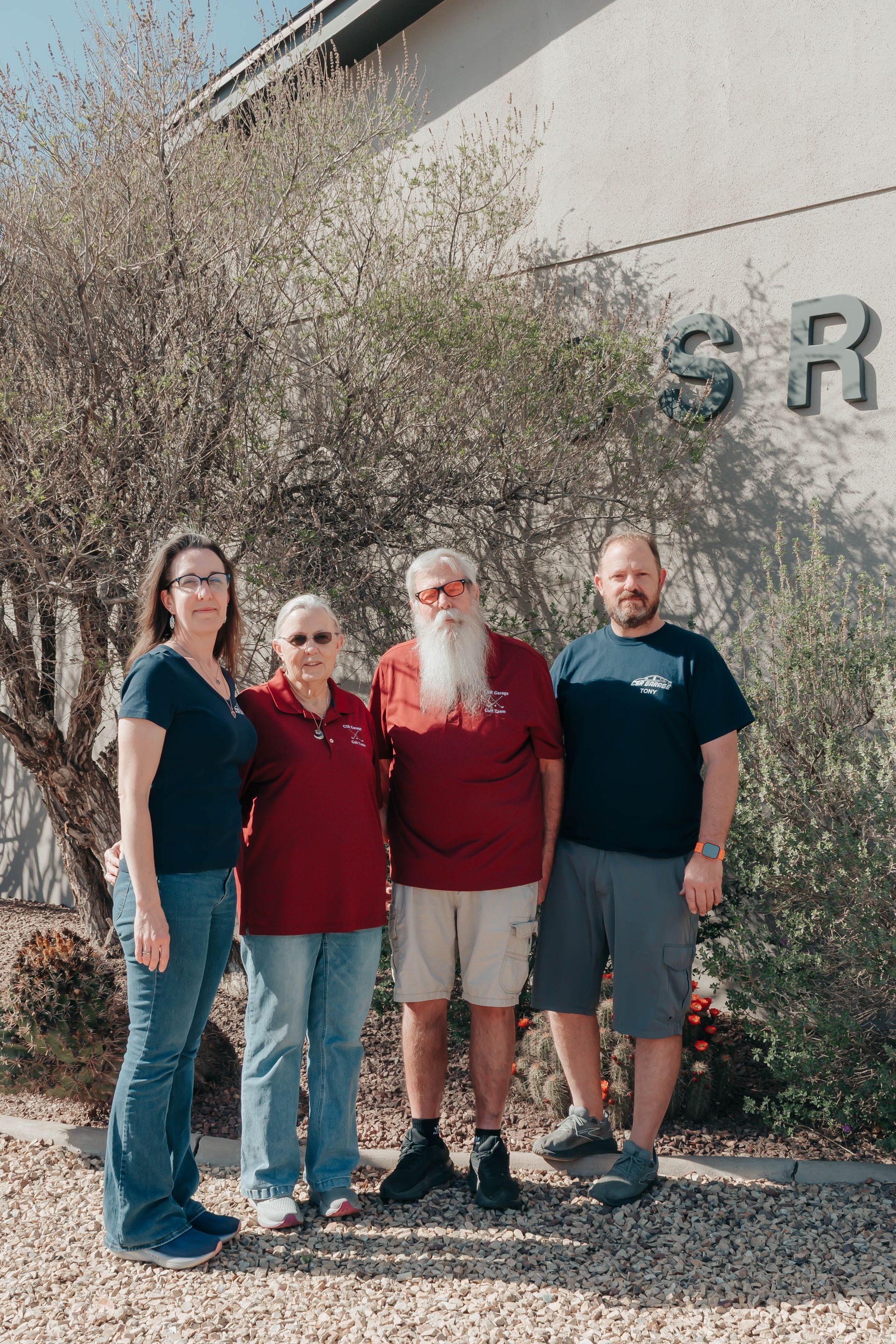 Four people standing outdoors beside a building, with bushes and partial wall lettering in the background.