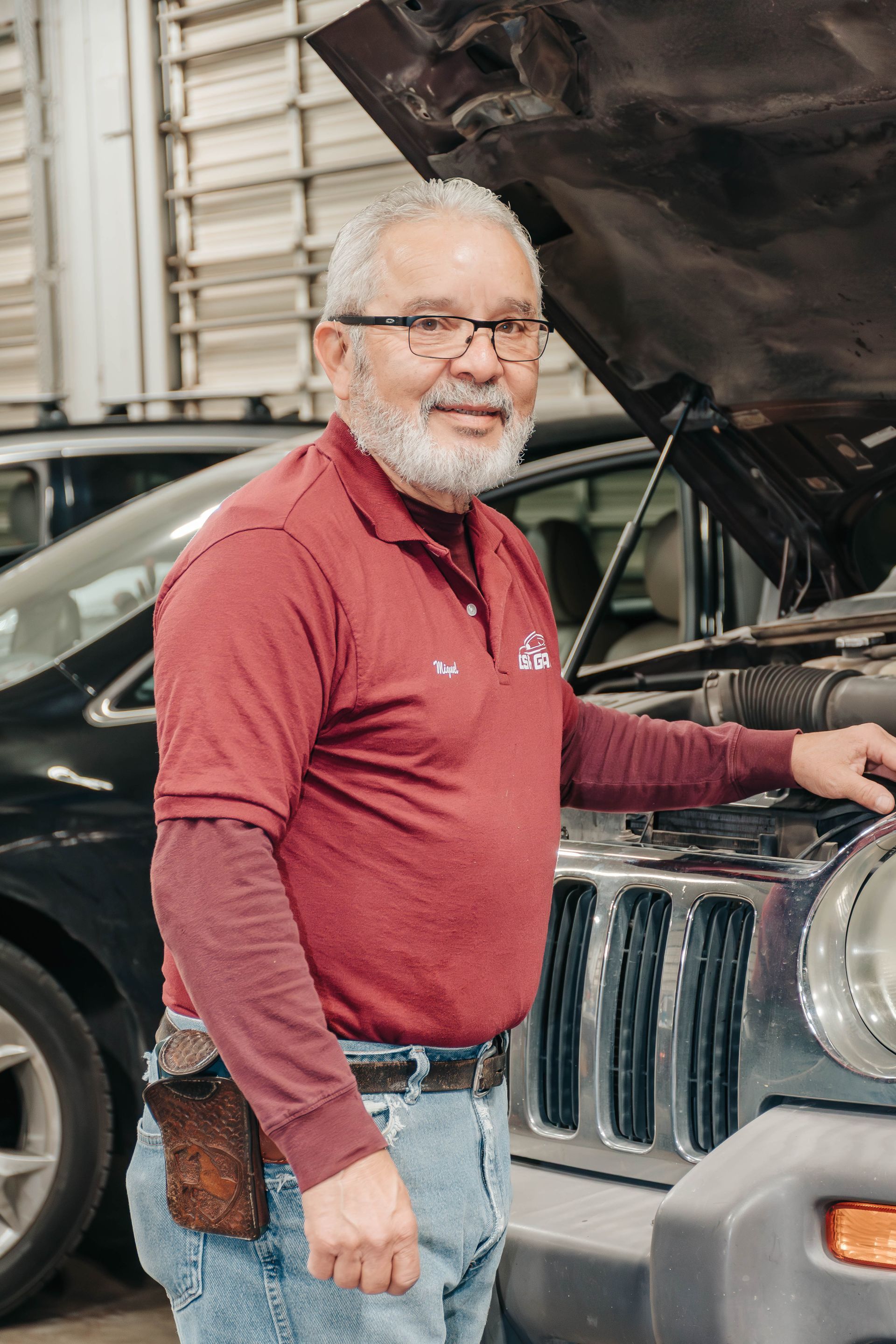 Man in red sweater standing by a car with its hood open, smiling in a garage