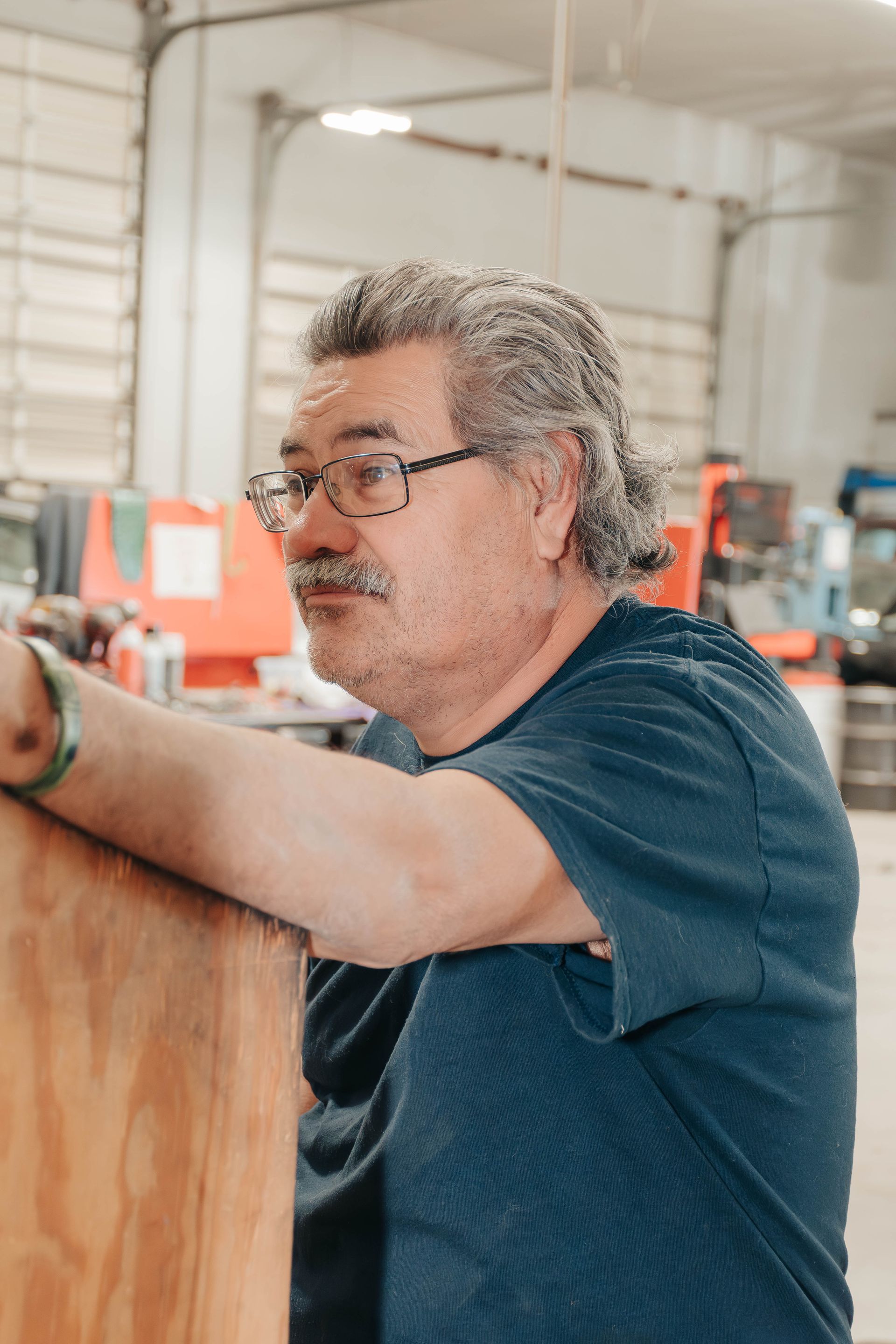 Man in blue shirt working in a workshop, leaning on a wooden surface and looking left