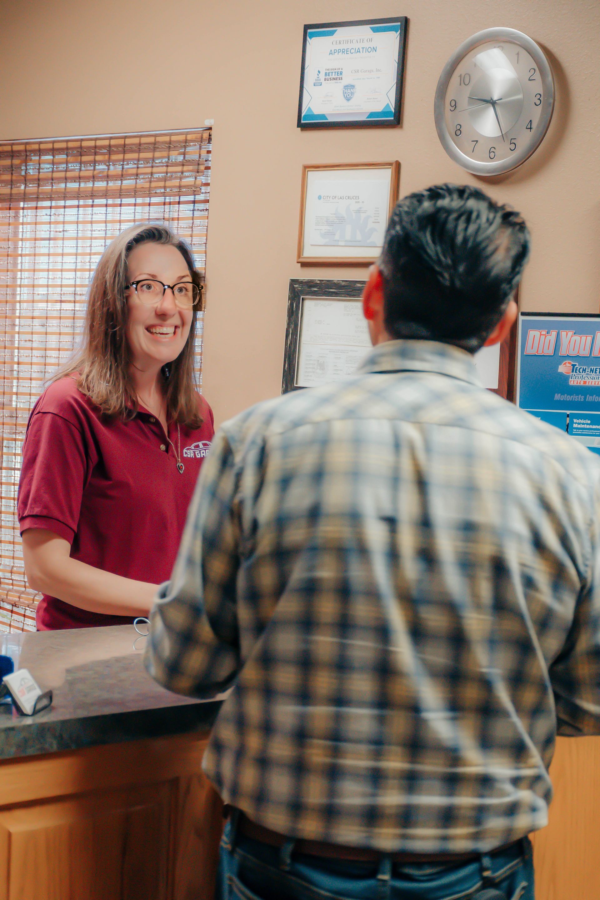 Woman at a reception desk talking to a customer in an office, with framed certificates and a wall clock.