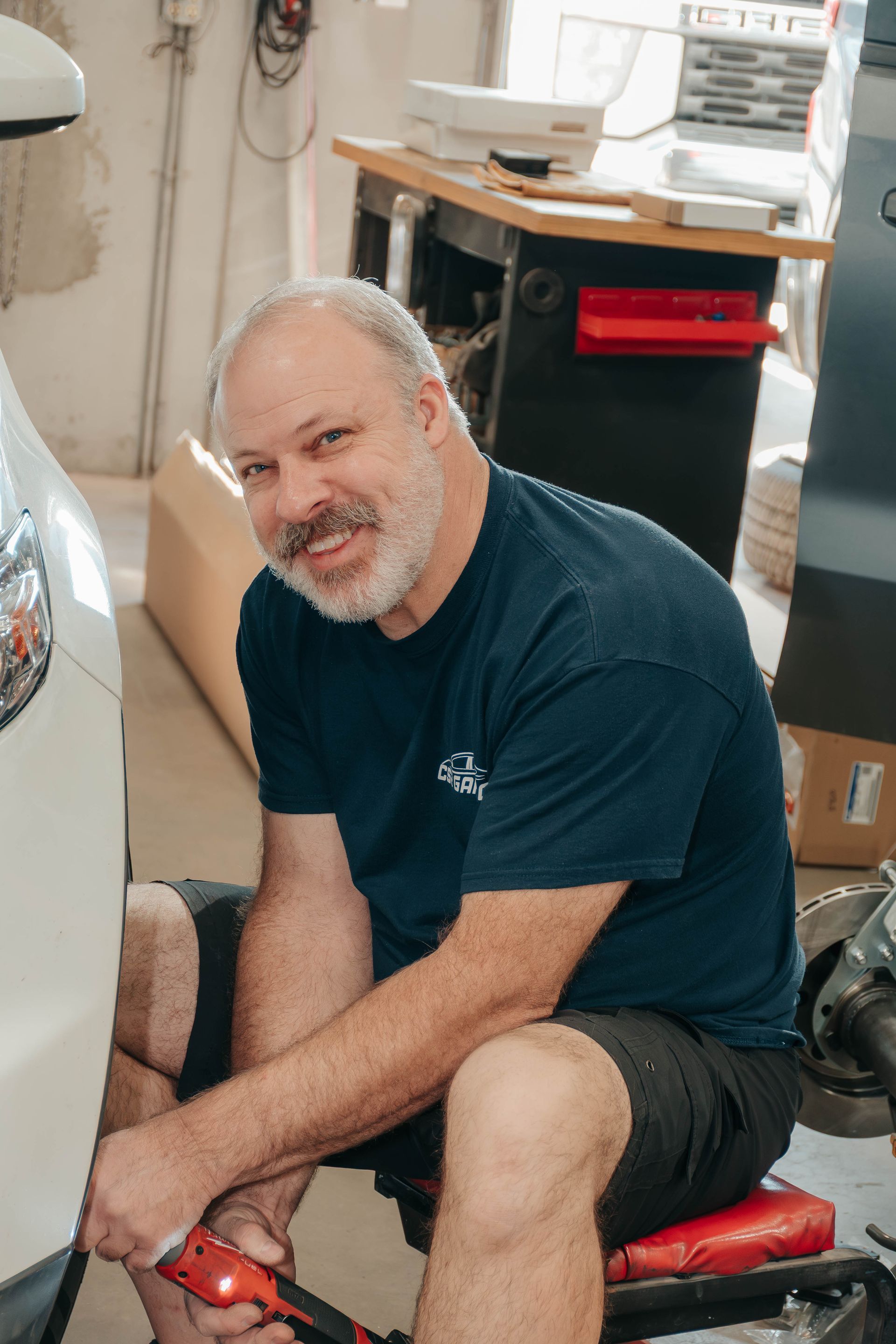 Man sitting on a red stool in a cluttered workspace, smiling at the camera.
