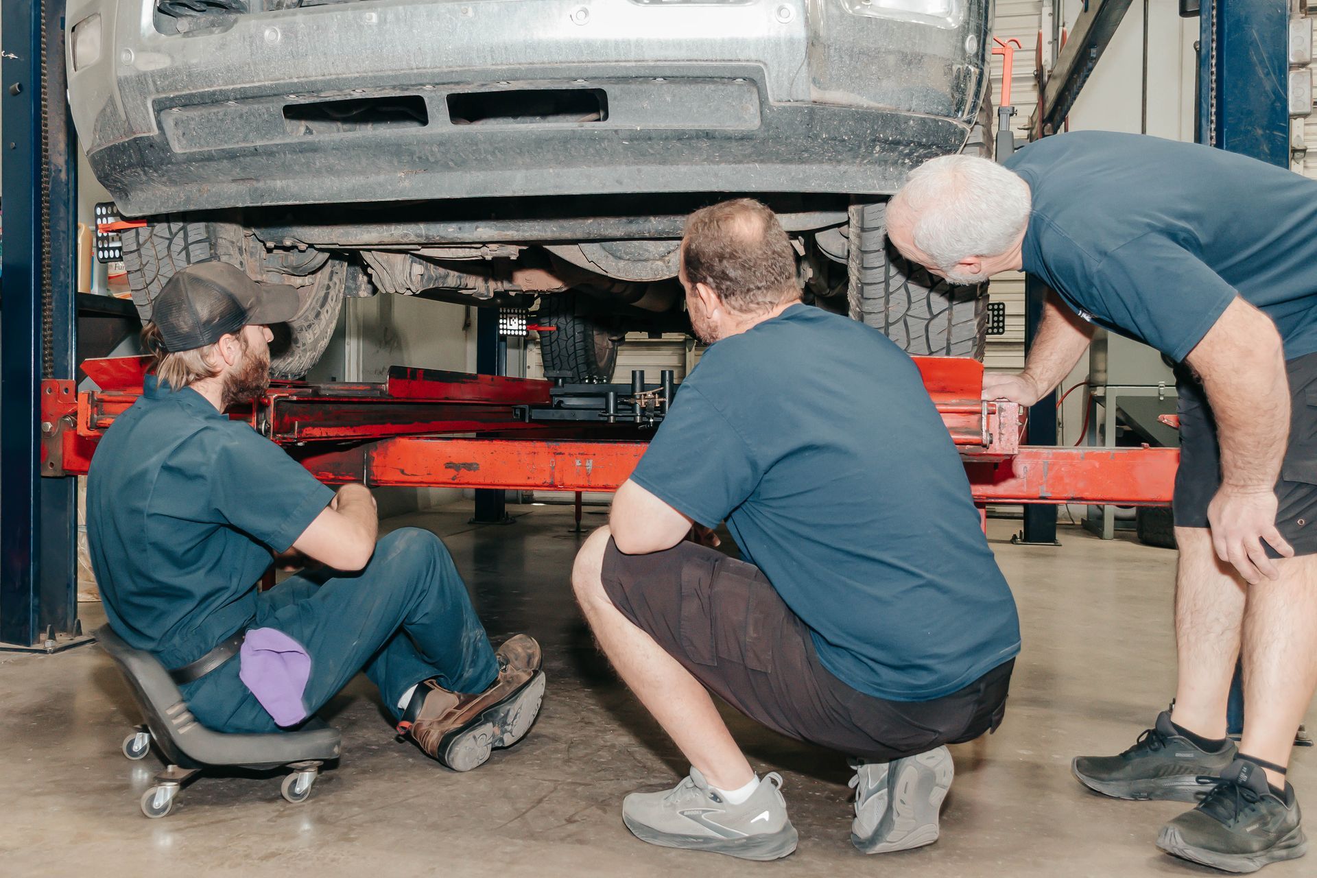 Three mechanics inspecting the underside of a car in a garage, with one crouched and two kneeling.