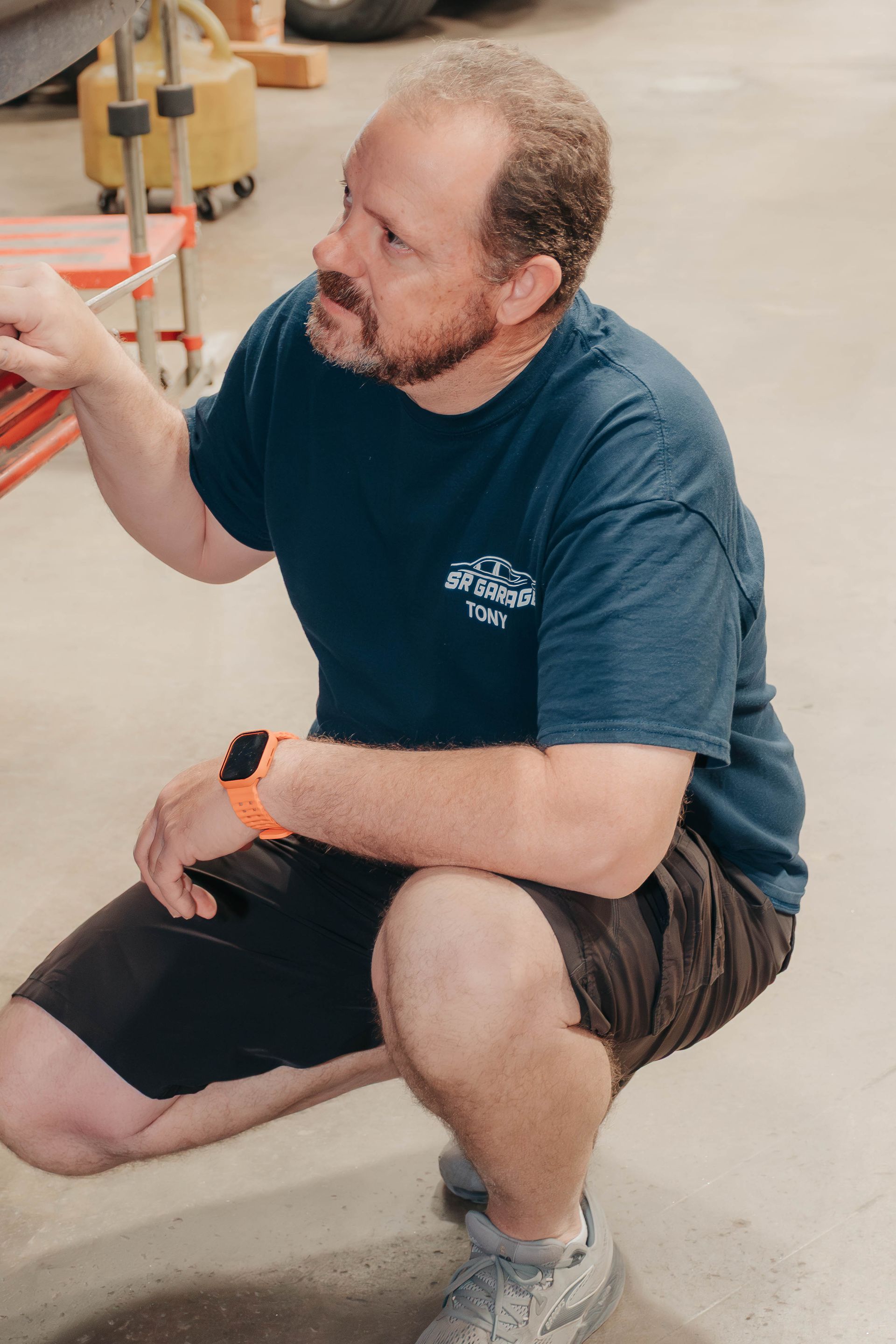 Kneeling man in a teal T-shirt and black shorts, gesturing in an indoor workshop.