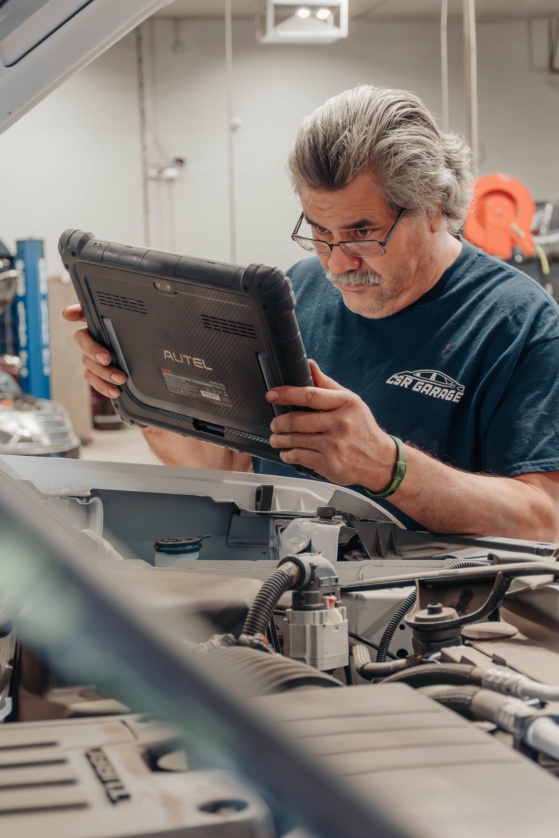 Mechanic in a blue coverall using a diagnostic tablet beside an open car hood in a workshop