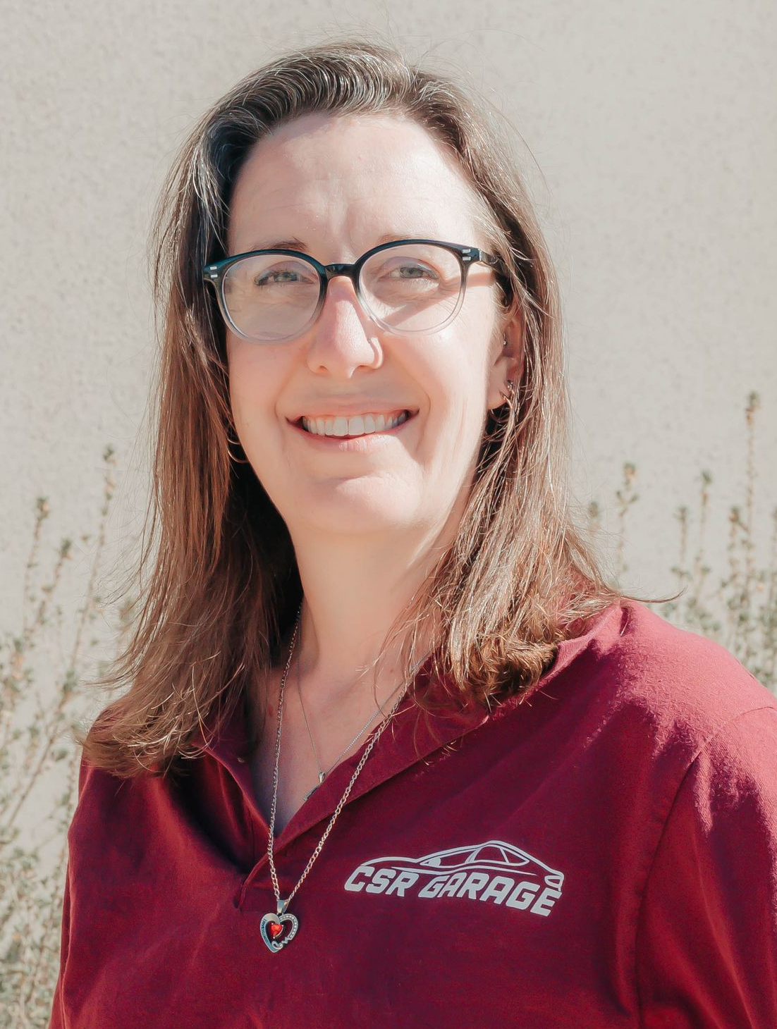 Smiling person with glasses wearing a maroon shirt and necklace, standing outdoors by a light wall.
