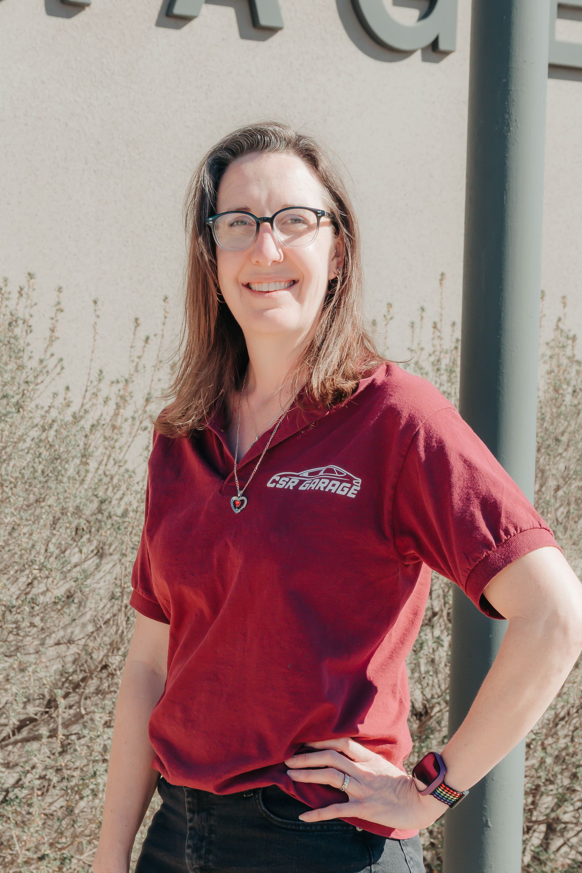 Woman in maroon shirt standing outdoors, smiling with hand on hip in front of a building.
