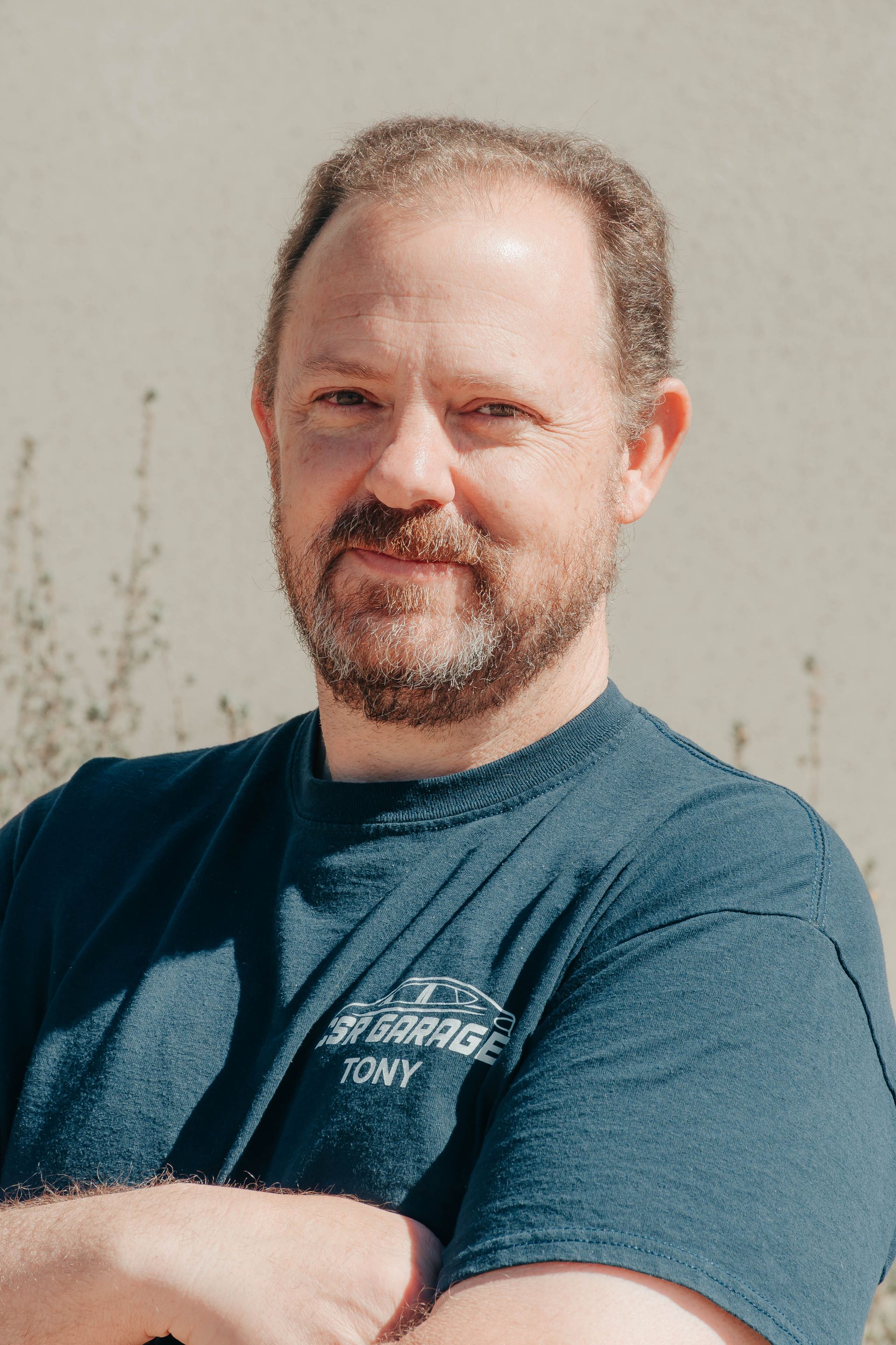 Man with a beard in a blue T-shirt, arms crossed, standing outdoors against a light wall.