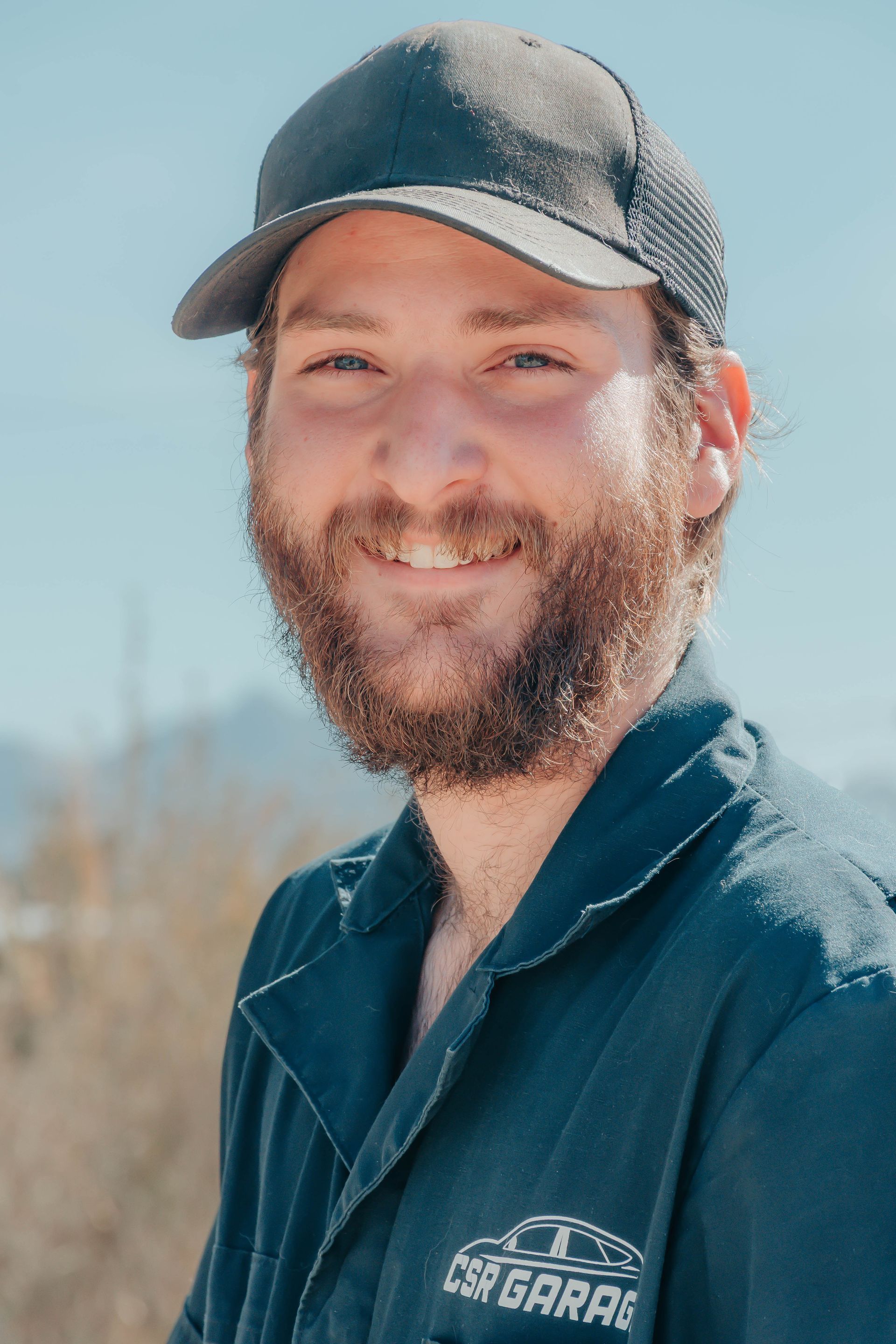 Smiling bearded man in a dark cap and shirt standing outdoors in sunlight