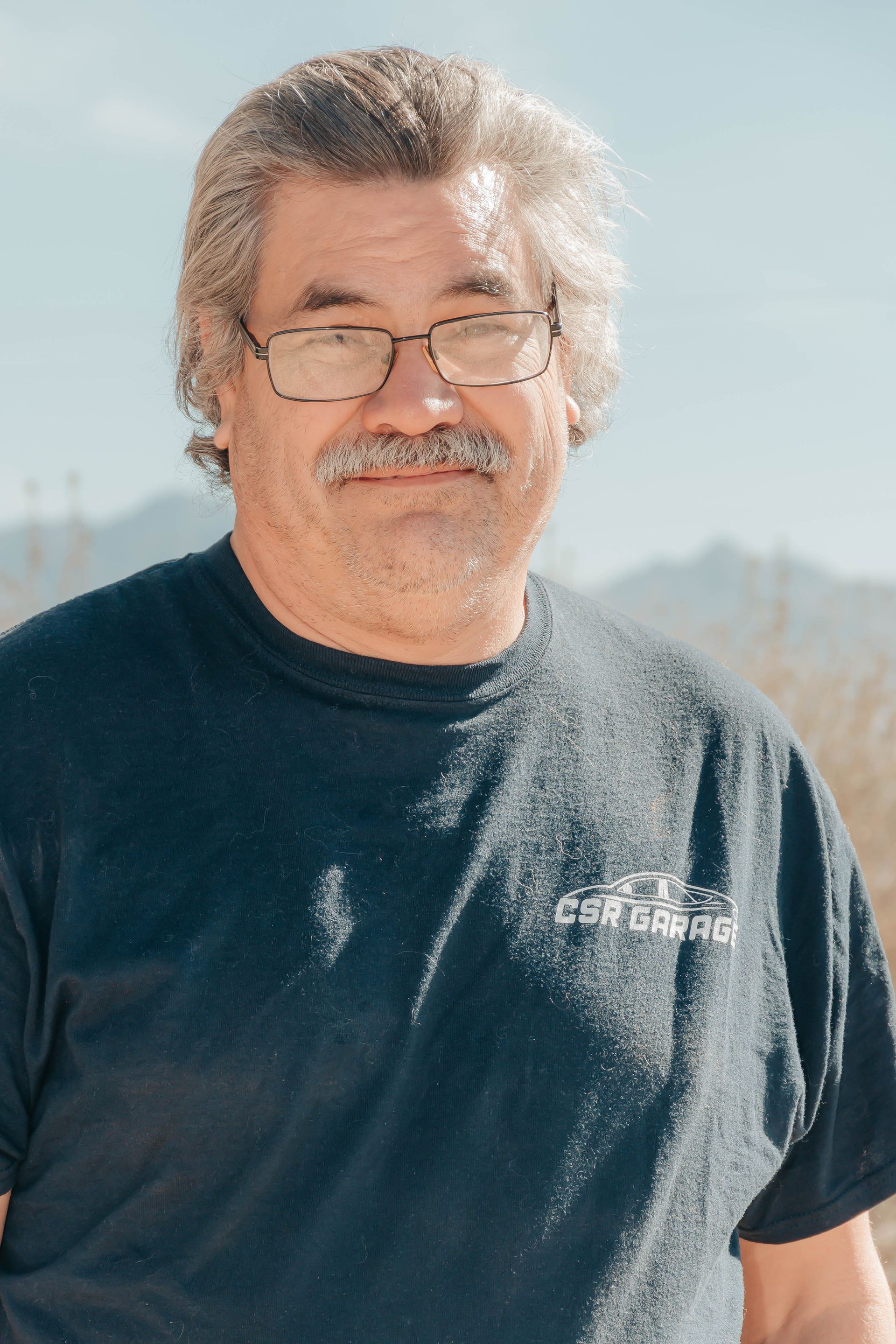 Man with glasses and mustache wearing a dark T-shirt outdoors in bright sunlight