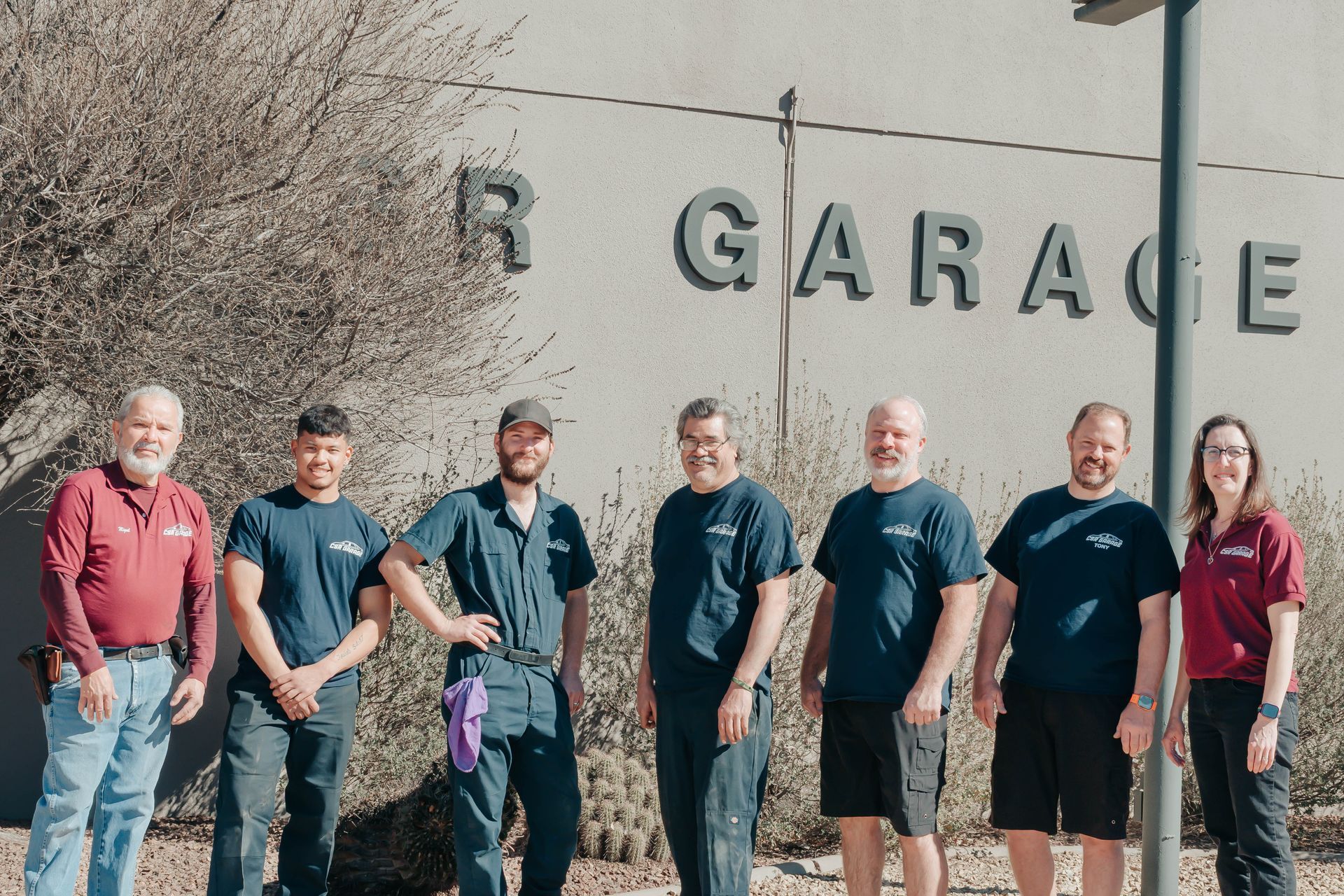 Seven people standing outdoors in front of a garage sign, posing together in casual clothing.