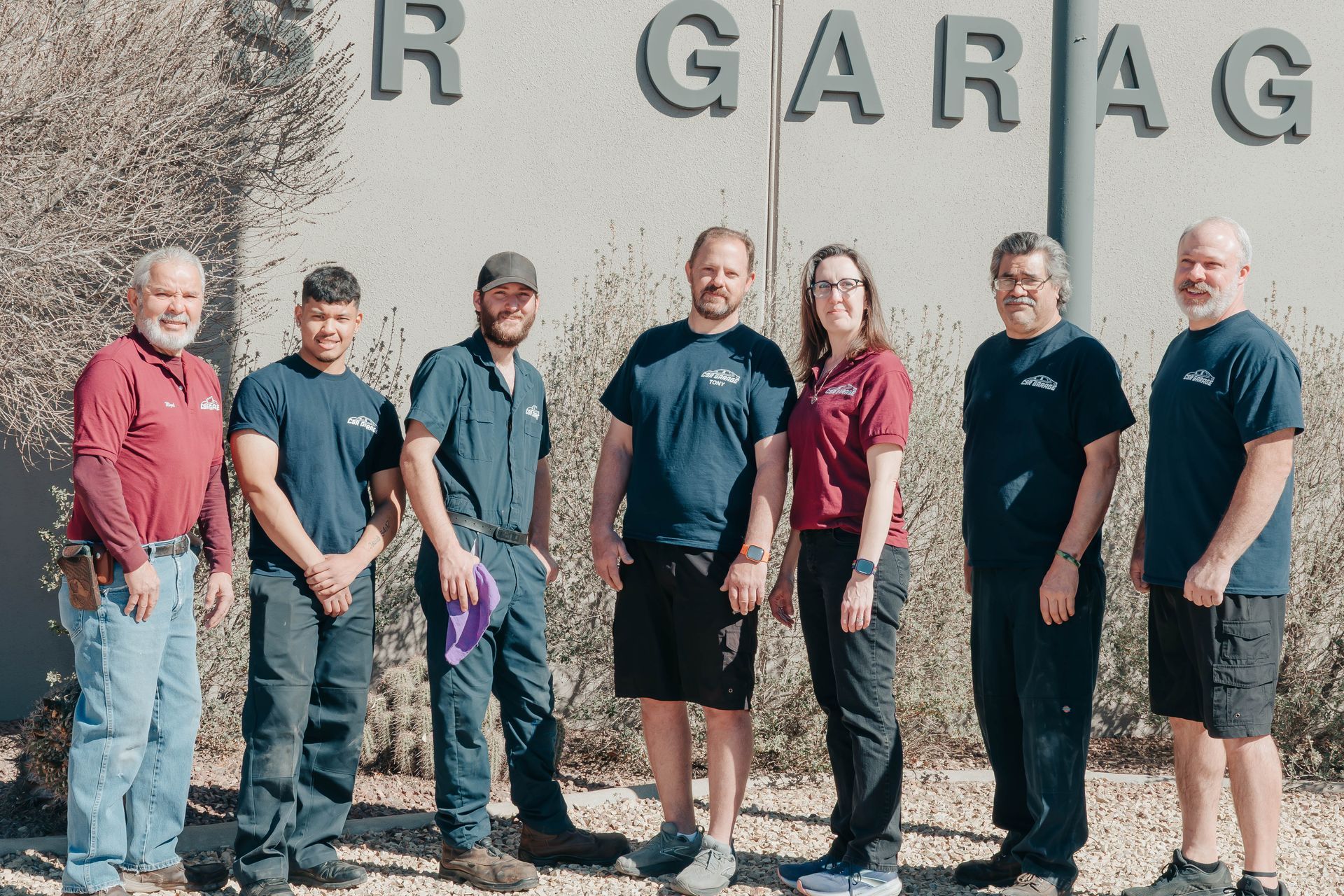 Six people standing outside a garage with a stone wall and large letters behind them