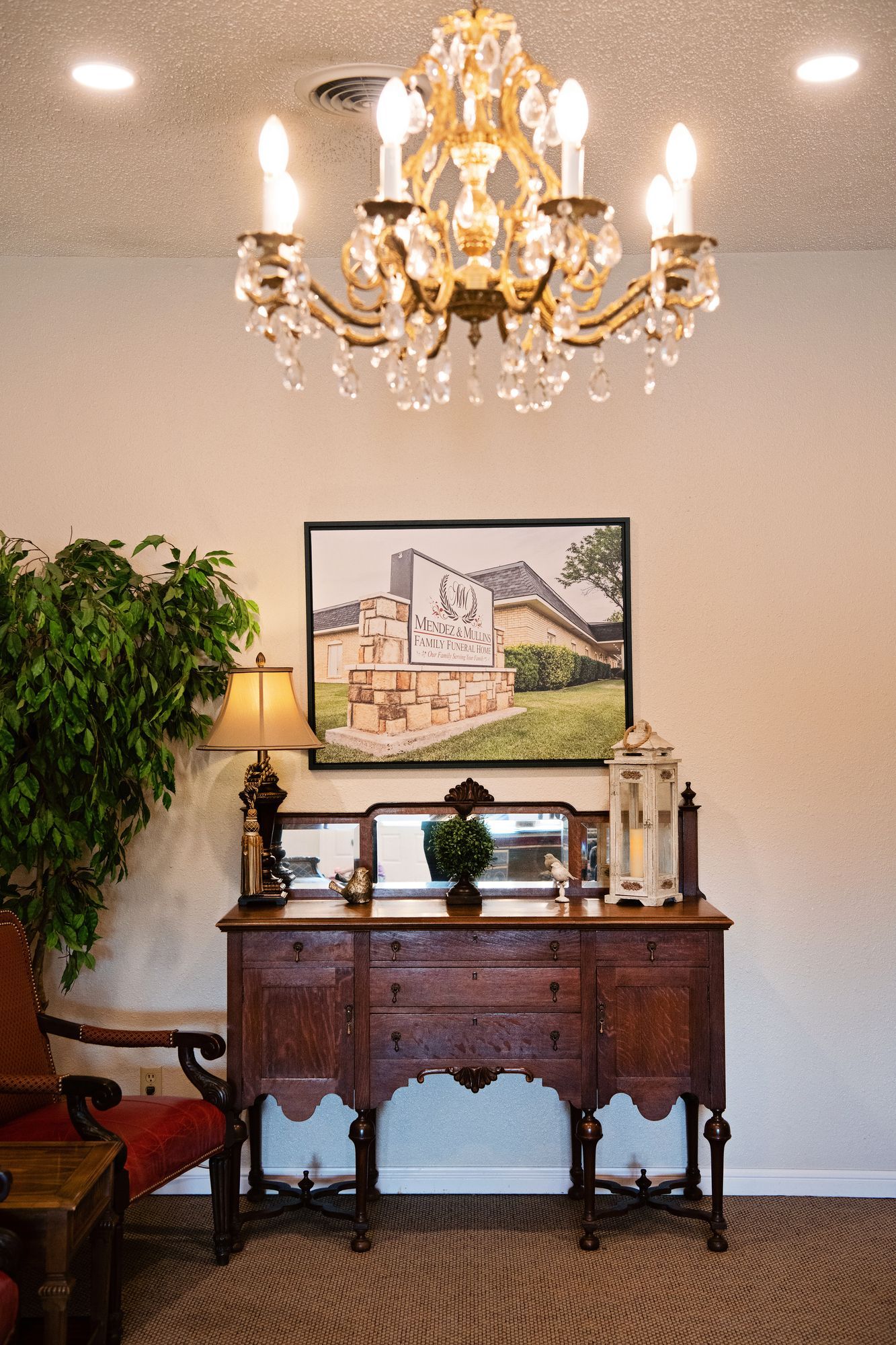 Elegant foyer with chandelier, ornate wooden table, framed art, lamp, and potted plants against a beige wall.