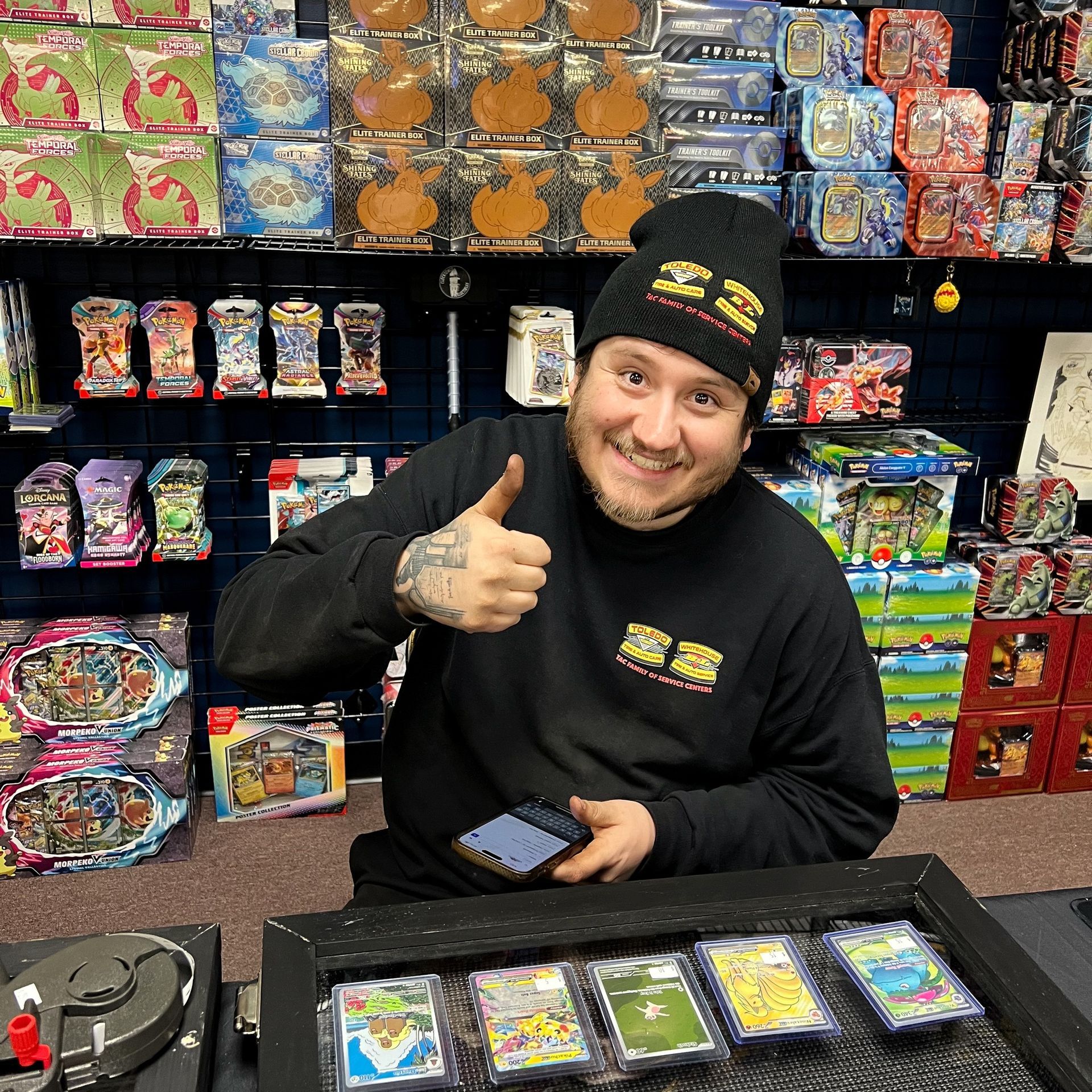 Man in beanie and black shirt, thumbs up, surrounded by collectible card game products.