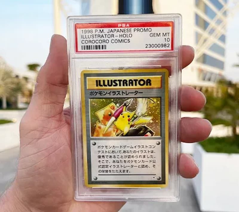 A hand holds a PSA 10 graded Japanese Pokémon Illustrator promo card in a plastic slab against a blurred outdoor backdrop.