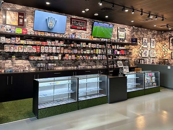 Inside a sports card store, shelves are stocked with cards. Display cases in front. TVs and framed items on the wall.