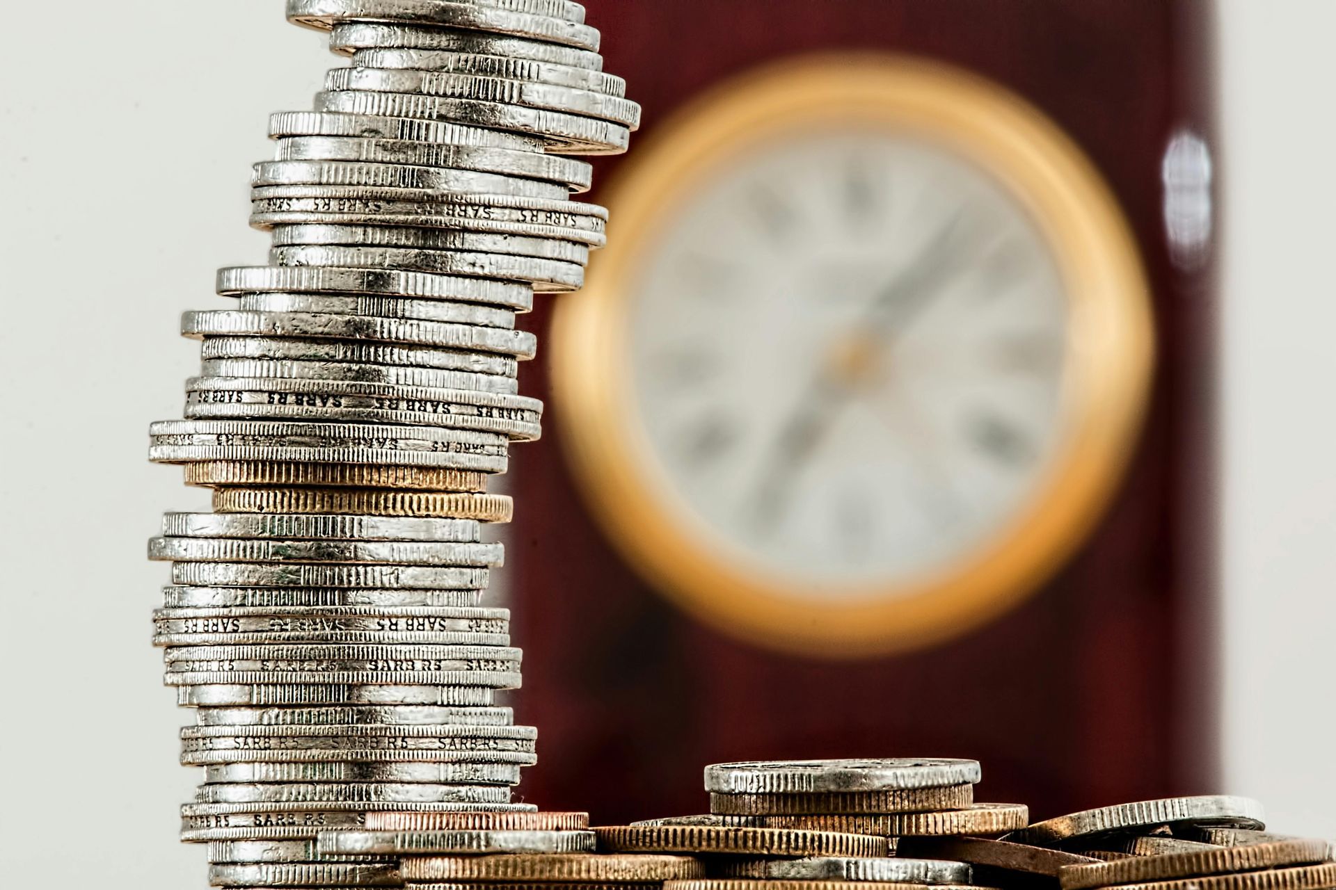 A tall, slightly tilted stack of silver coins sits in the foreground, with a blurred analog clock in the background.
