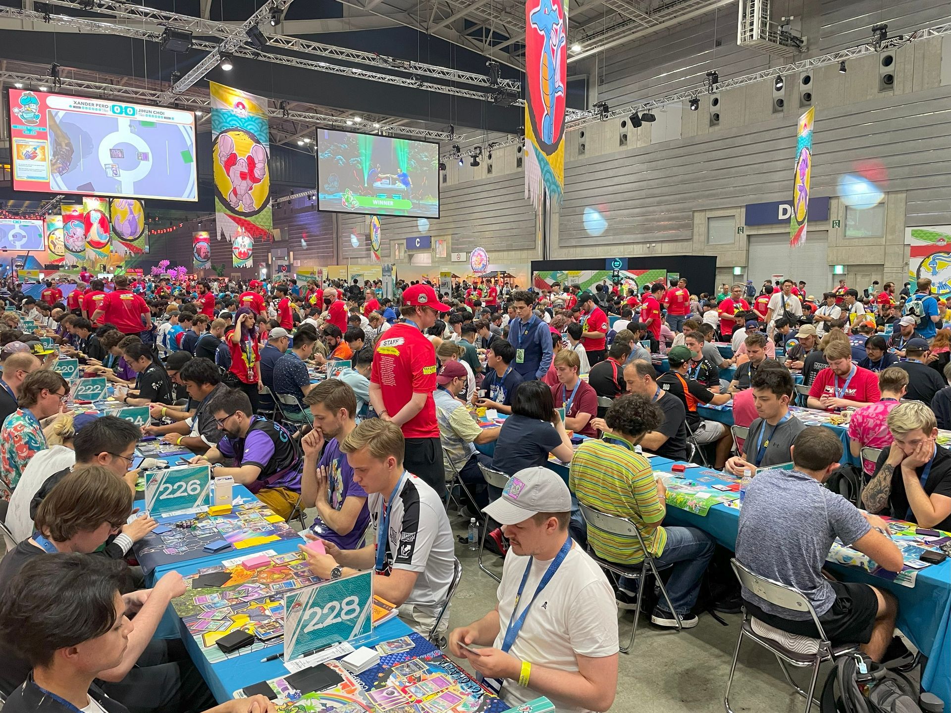 Large convention hall filled with people at tables, many playing card games. Bright lights, colorful banners, screens.