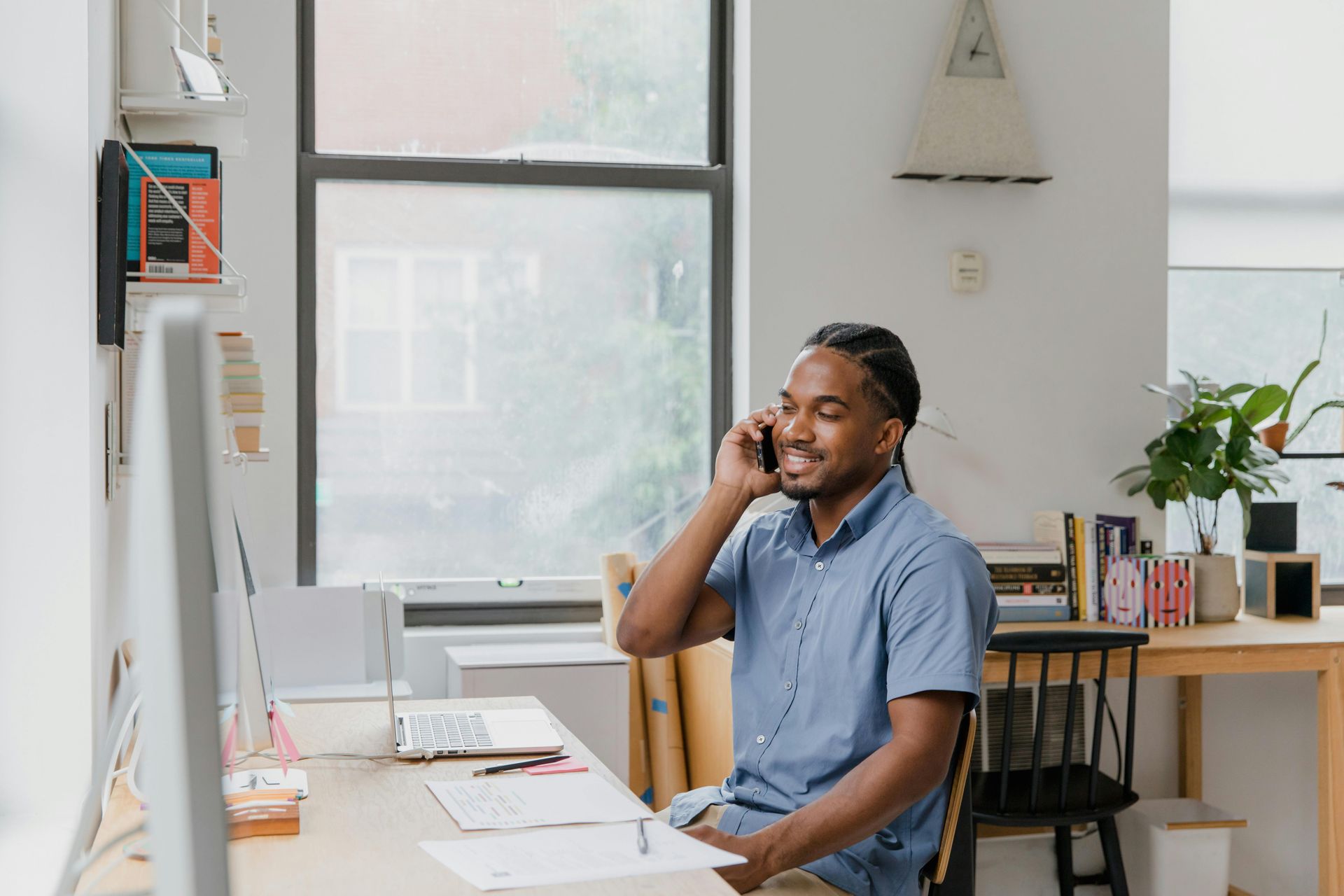 A person in a blue shirt smiling while talking on a phone at a desk in a home office.