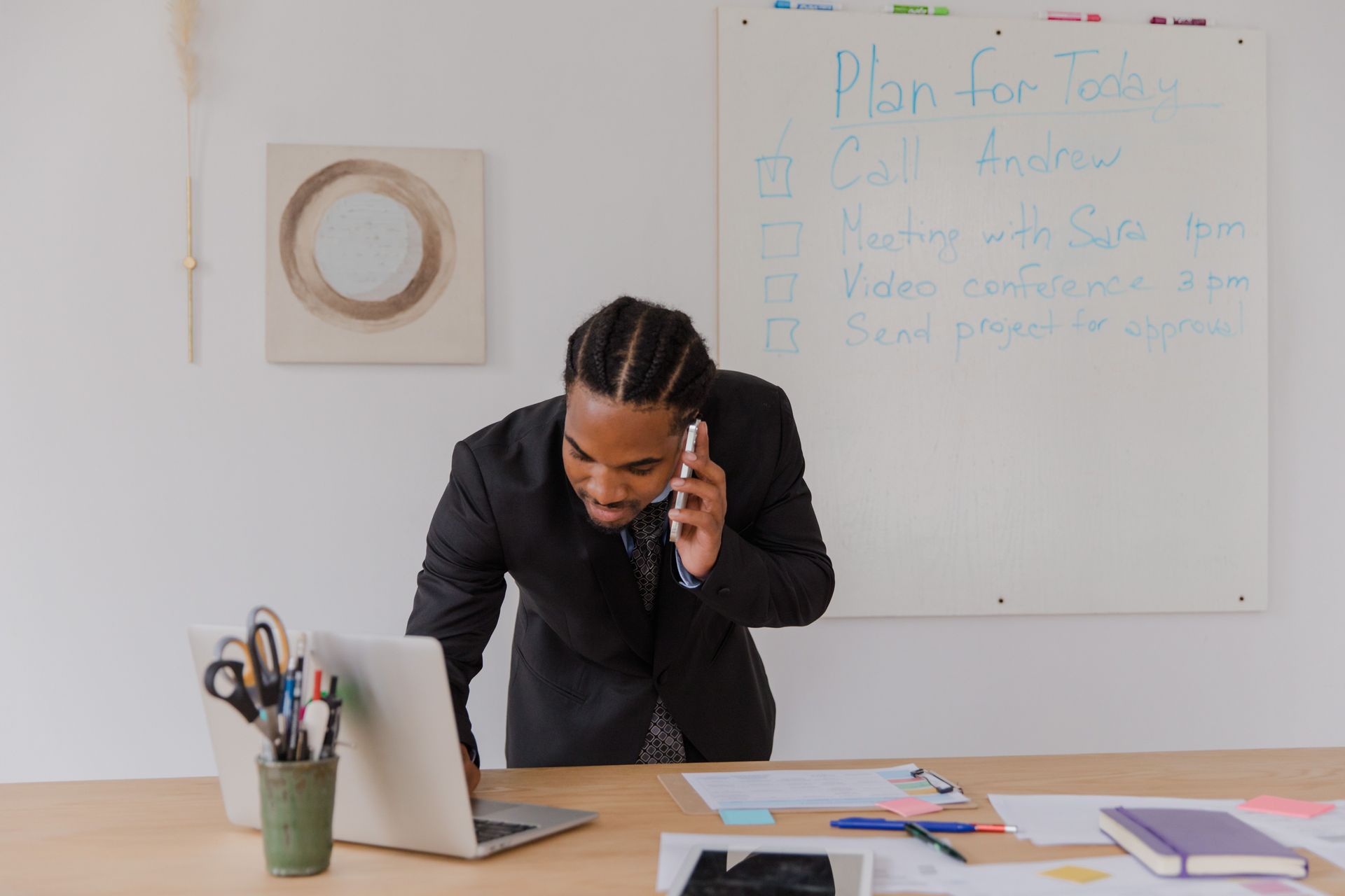 Person on a phone call at an office desk, leaning over a laptop with notes on a whiteboard behind.