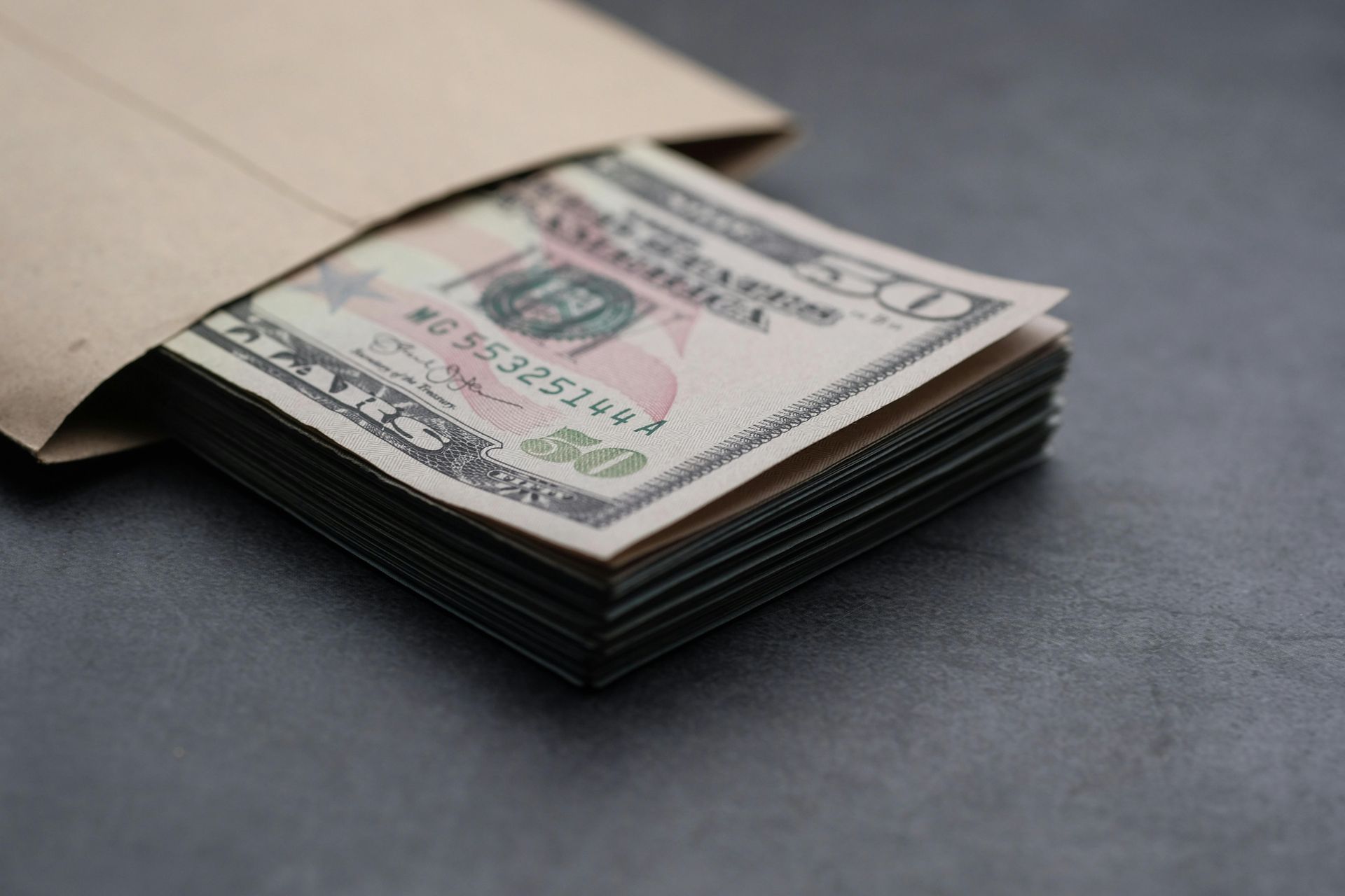 A stack of US fifty-dollar bills partially tucked inside a tan paper envelope on a dark gray surface.