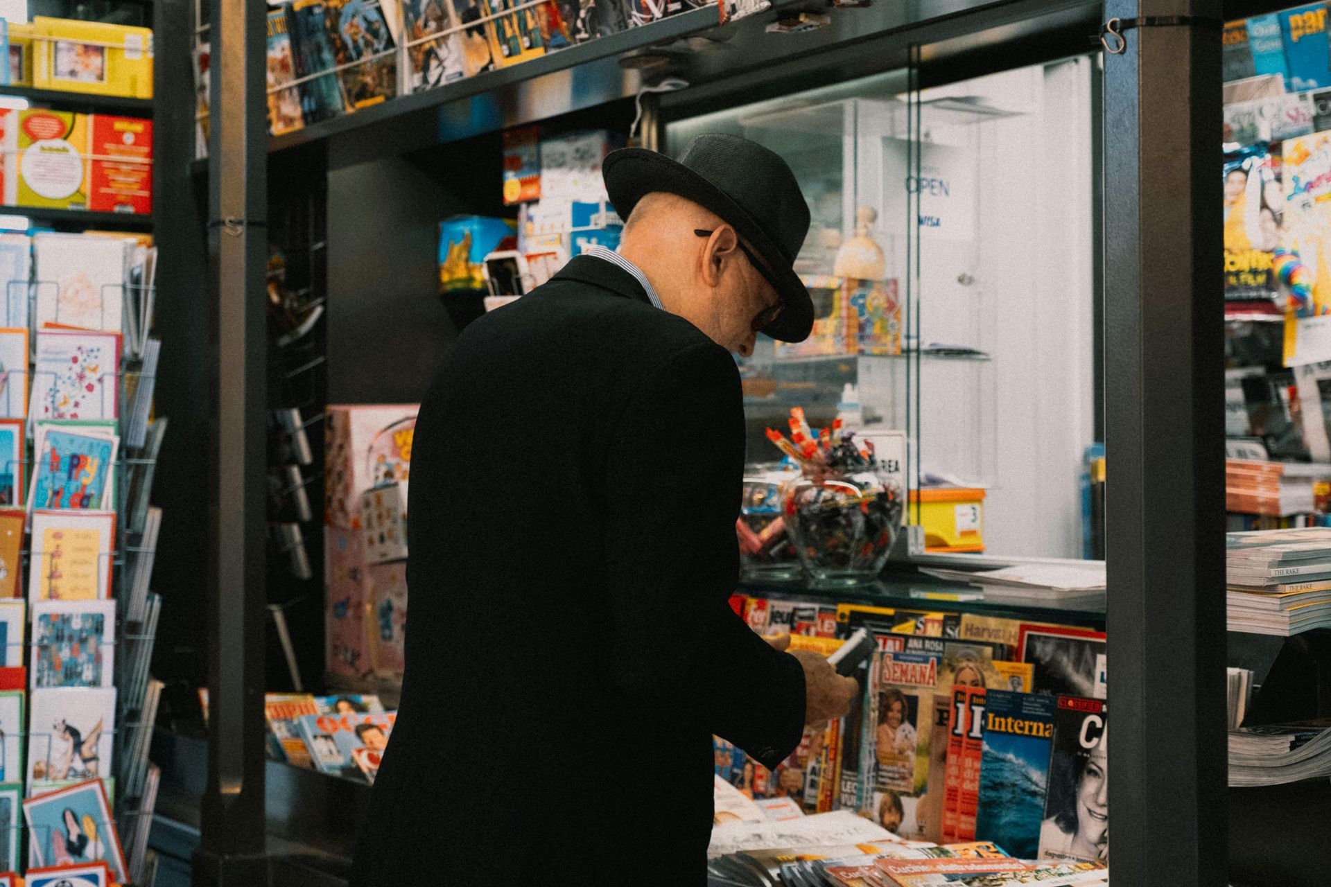 A person in a black coat and hat stands at a shop counter browsing through a selection of magazines and merchandise.