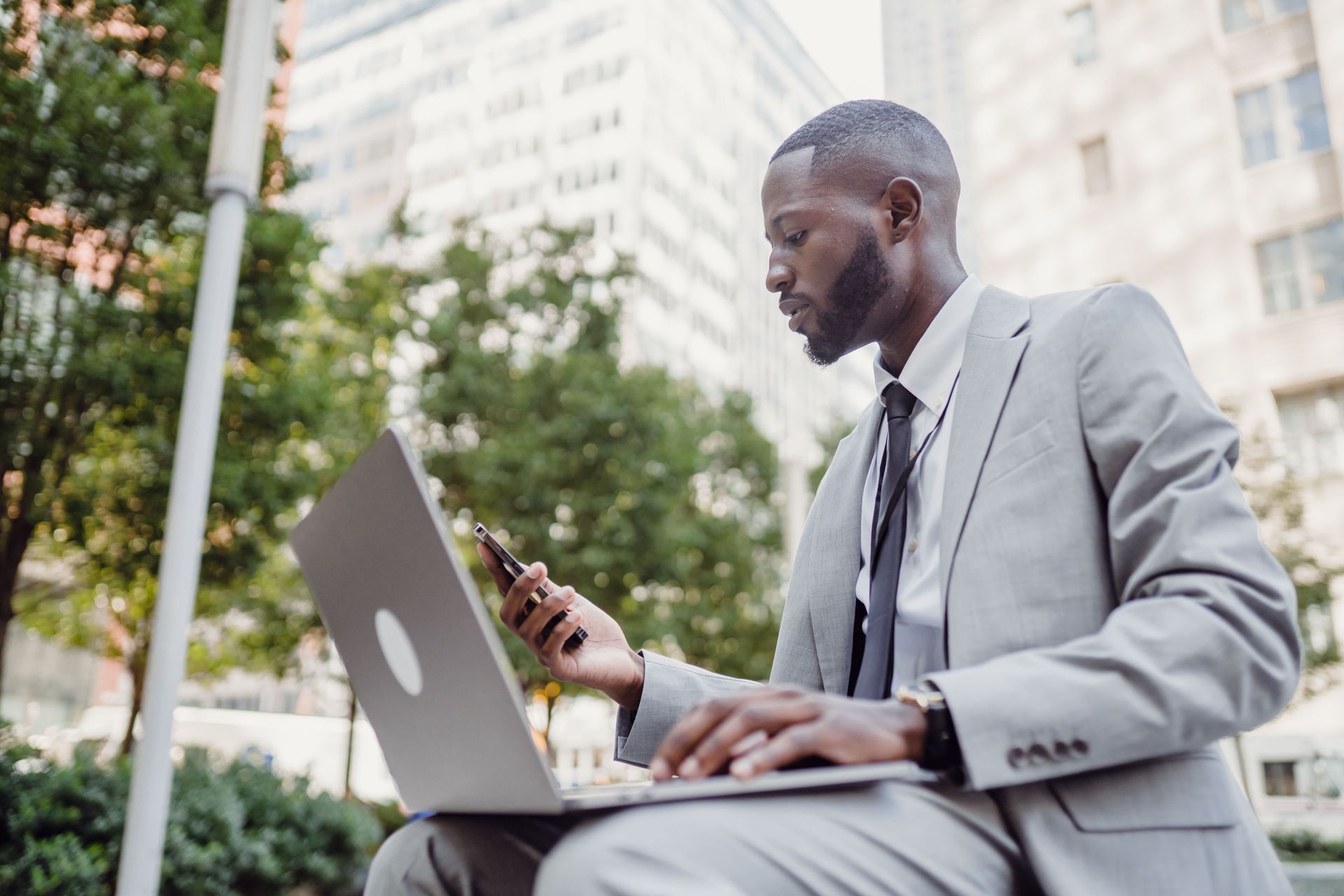 Man in a light gray suit using a laptop and phone outdoors in a city plaza