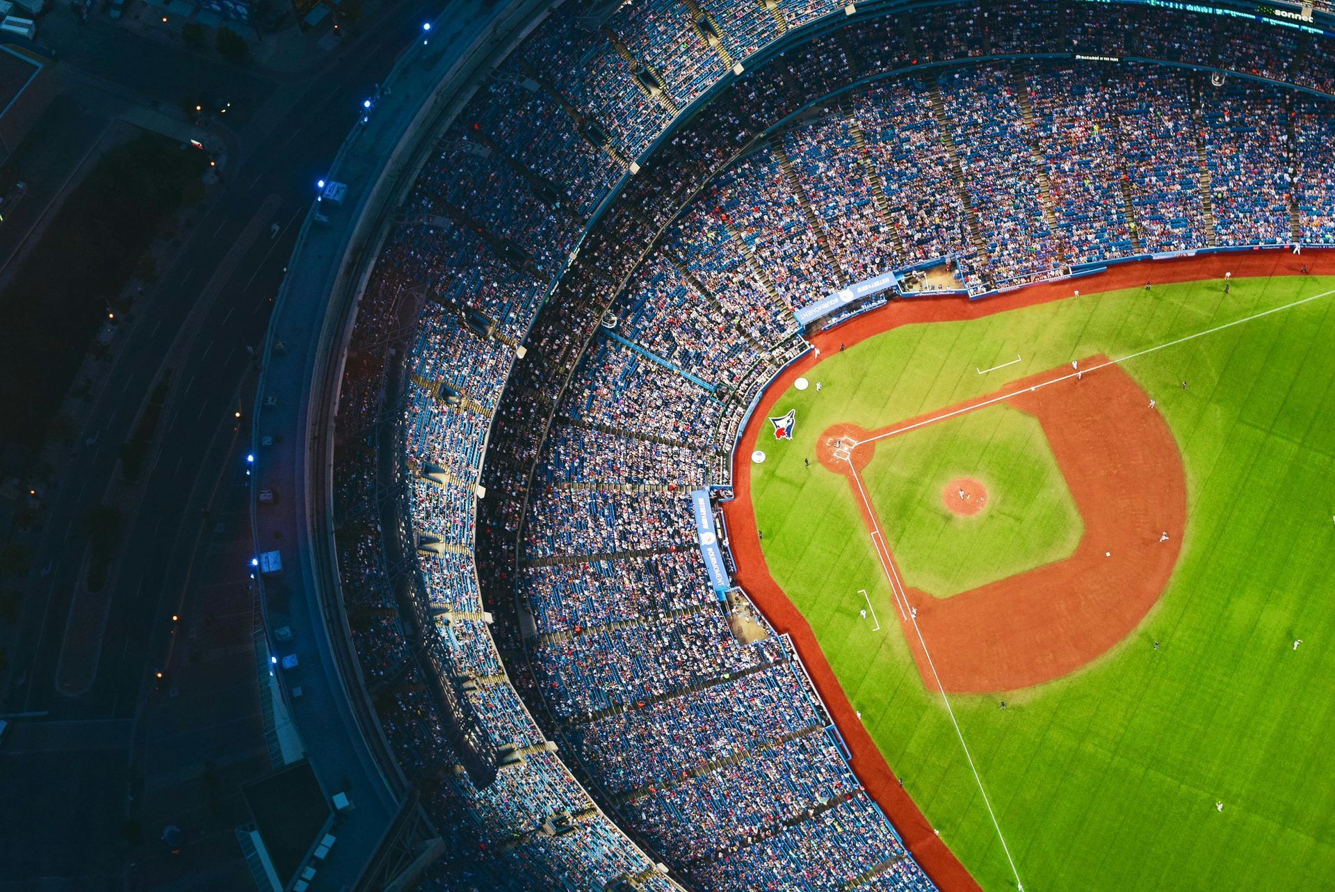 Overhead view of a baseball stadium at night, packed with spectators, green field and red dirt.