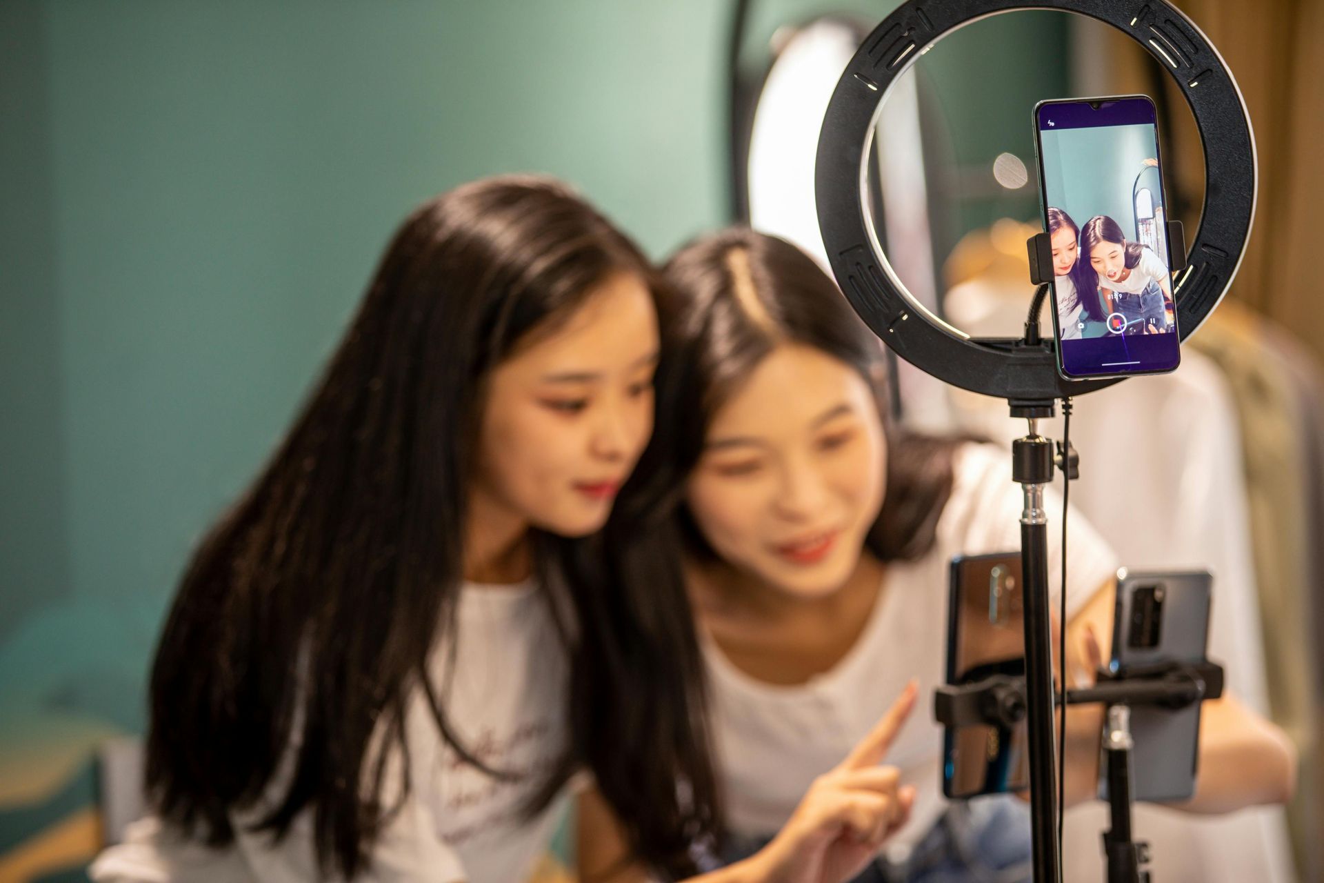 Two people recording with a phone and ring light. One points at a screen, both look at it closely.