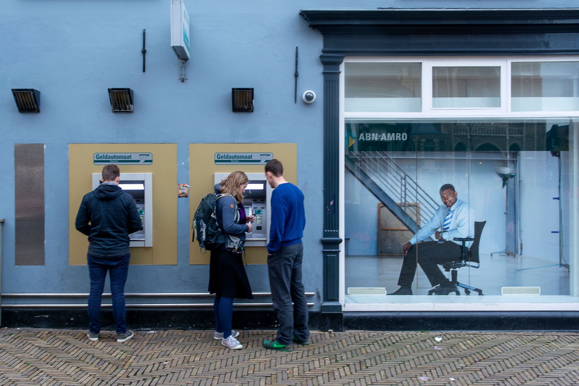 People standing at a wall-mounted display outside a blue building, with a seated man visible through the window.
