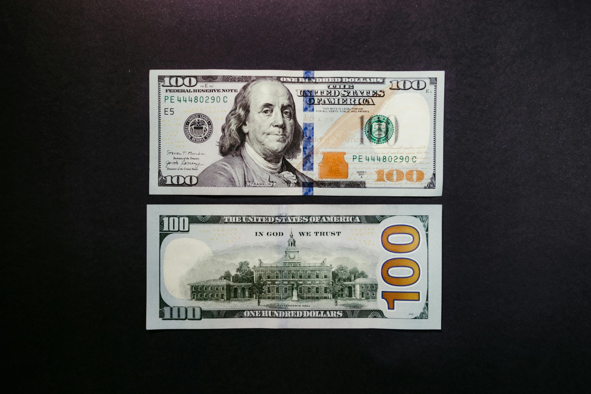 A U.S. $100 bill, face-up showing Benjamin Franklin, and reverse displaying the U.S. Treasury building.