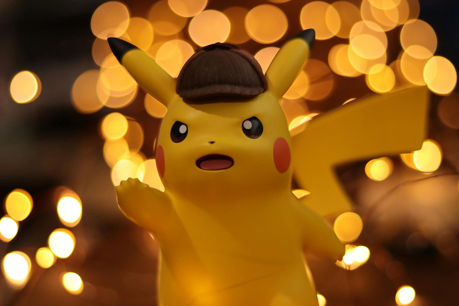 Pikachu wearing a brown detective hat, gesturing with a hand; background of golden bokeh lights.