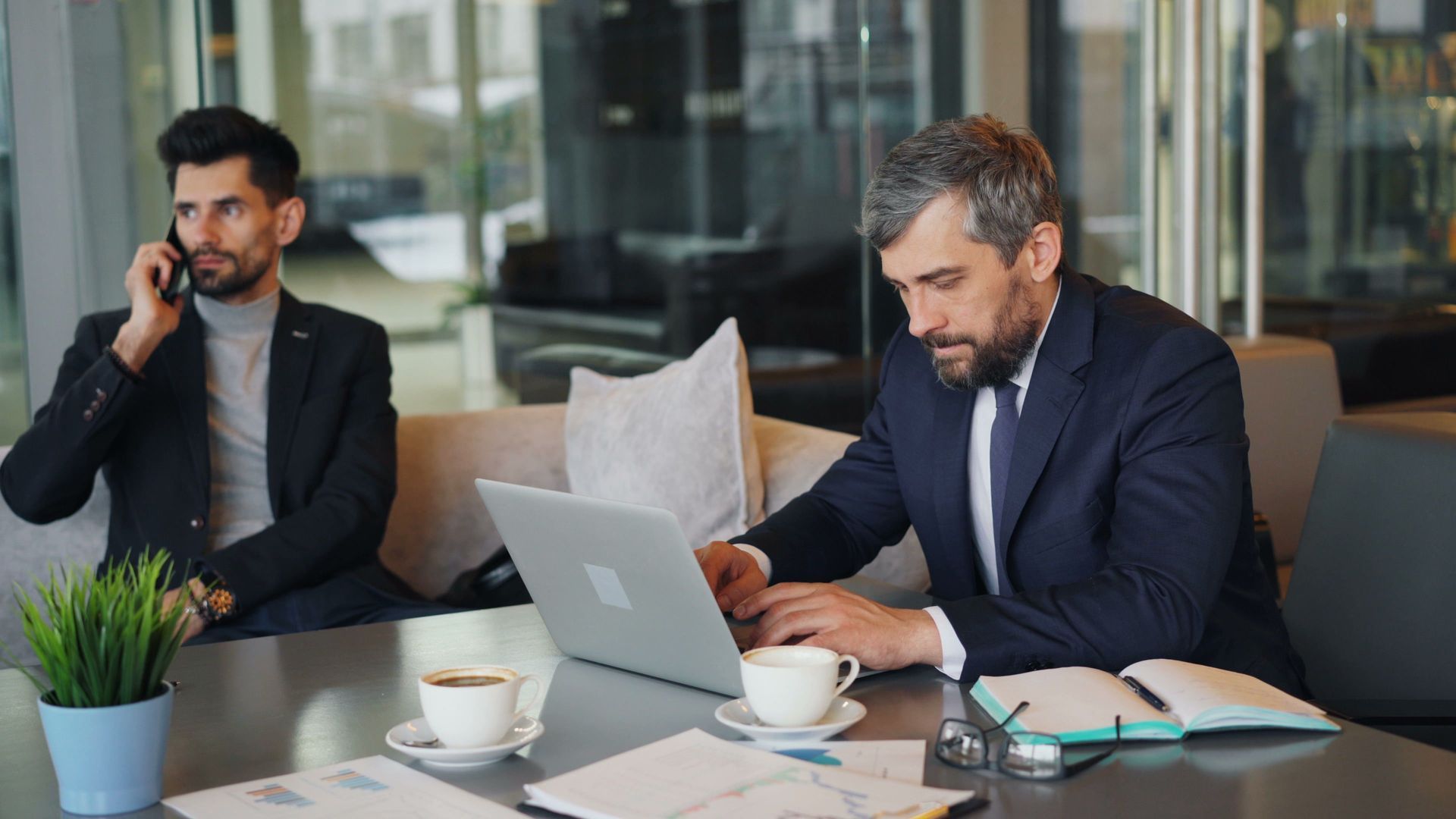 Two men in a modern office, one on the phone and one typing on a laptop at a table with coffee cups.