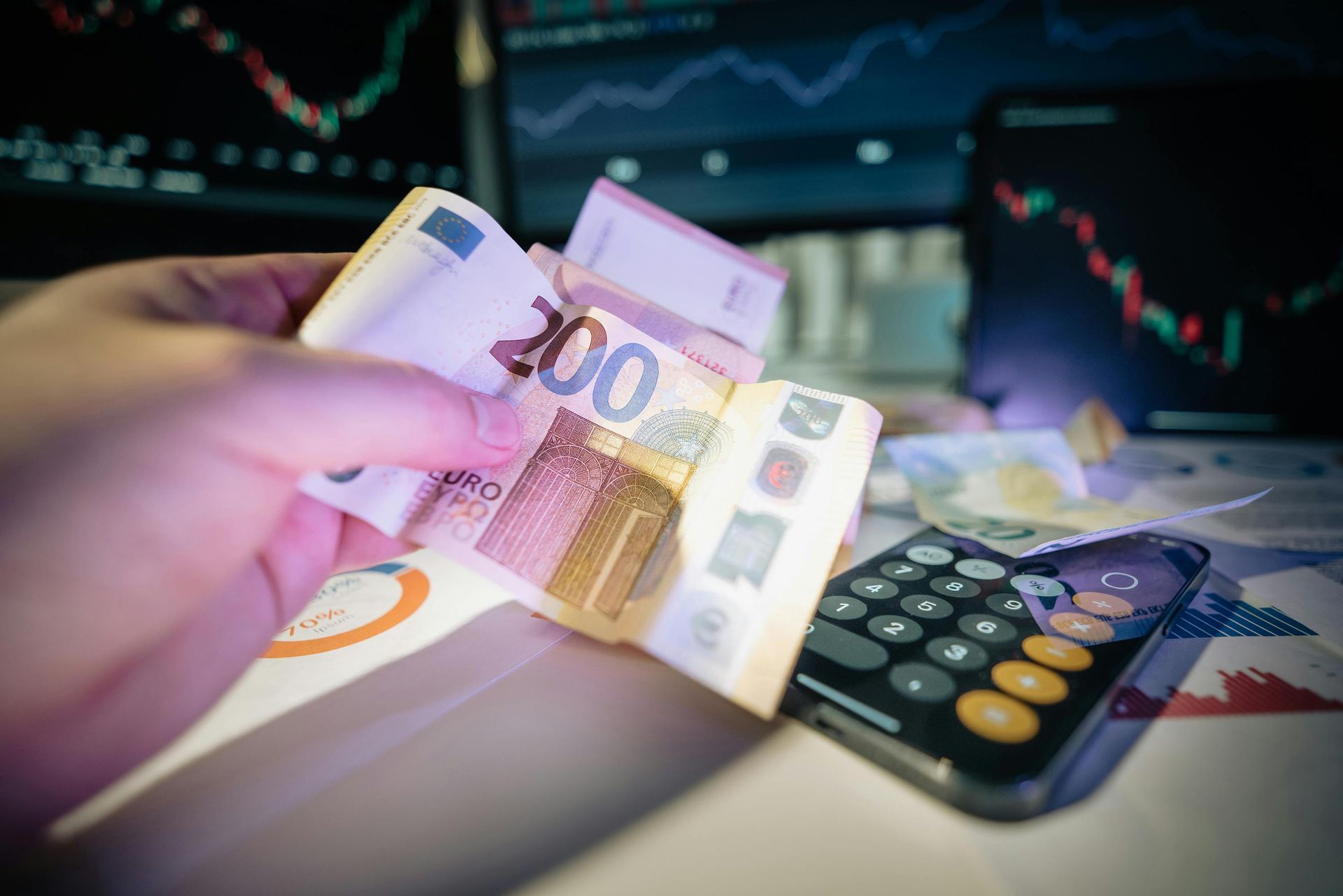 Hand holding Euro bills, with calculator, charts, and stock graphs in the background.