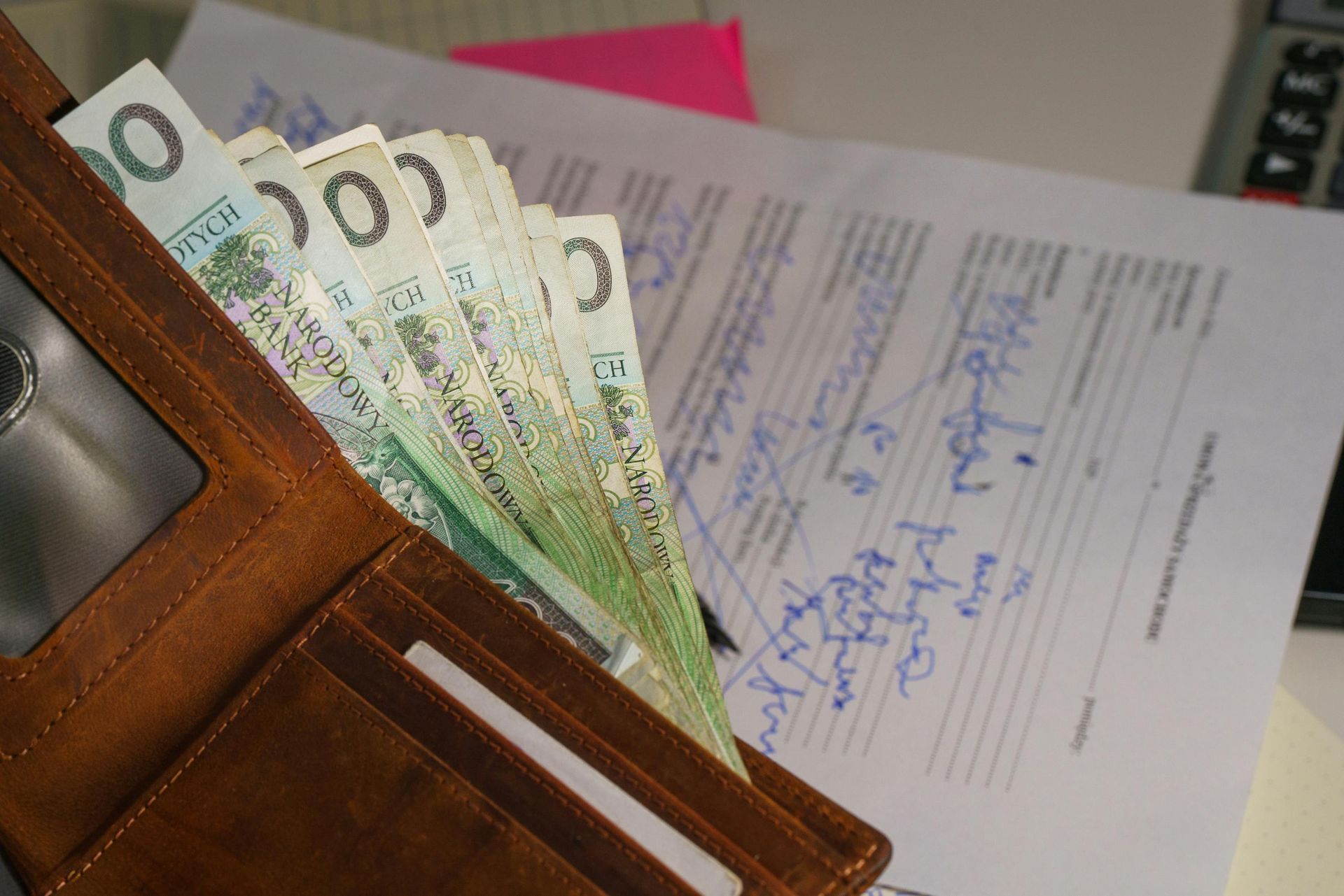 Brown leather wallet filled with Polish banknotes, resting on paperwork with signatures.