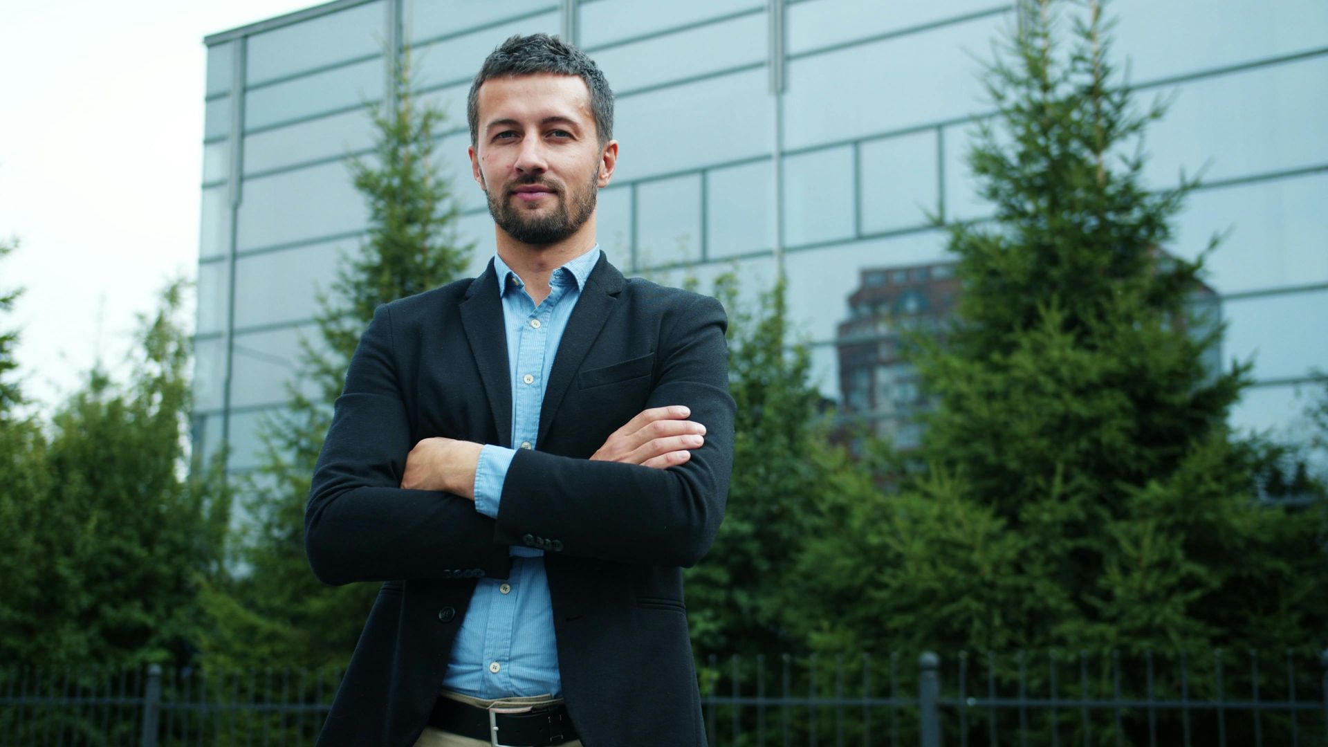 Man in a dark blazer stands with arms crossed outside a modern glass building.