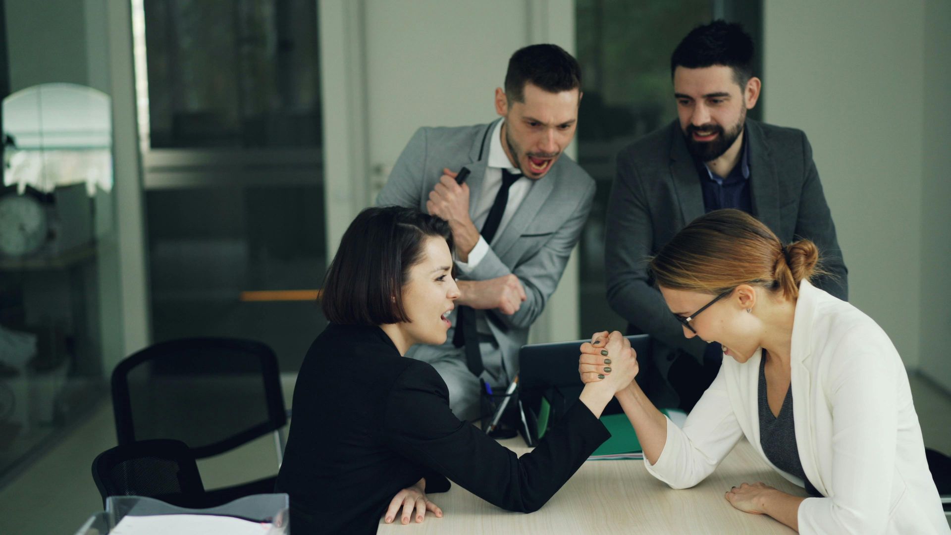 Colleagues in a meeting shaking hands over paperwork, with two others standing behind them in an office.