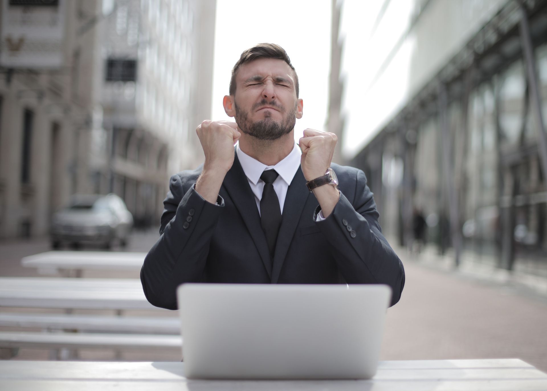Frustrated man in a suit at a laptop, fists raised in a city street setting