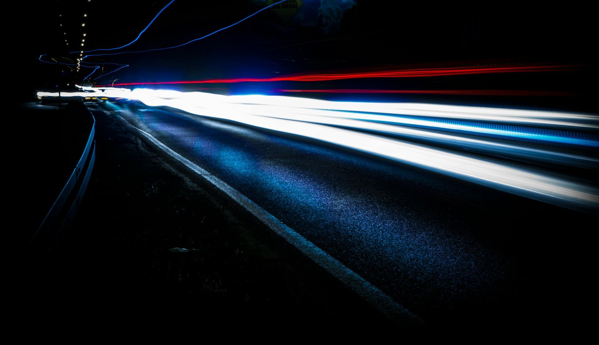 Long-exposure shot of white and red light trails from vehicles moving along a dark road at night.