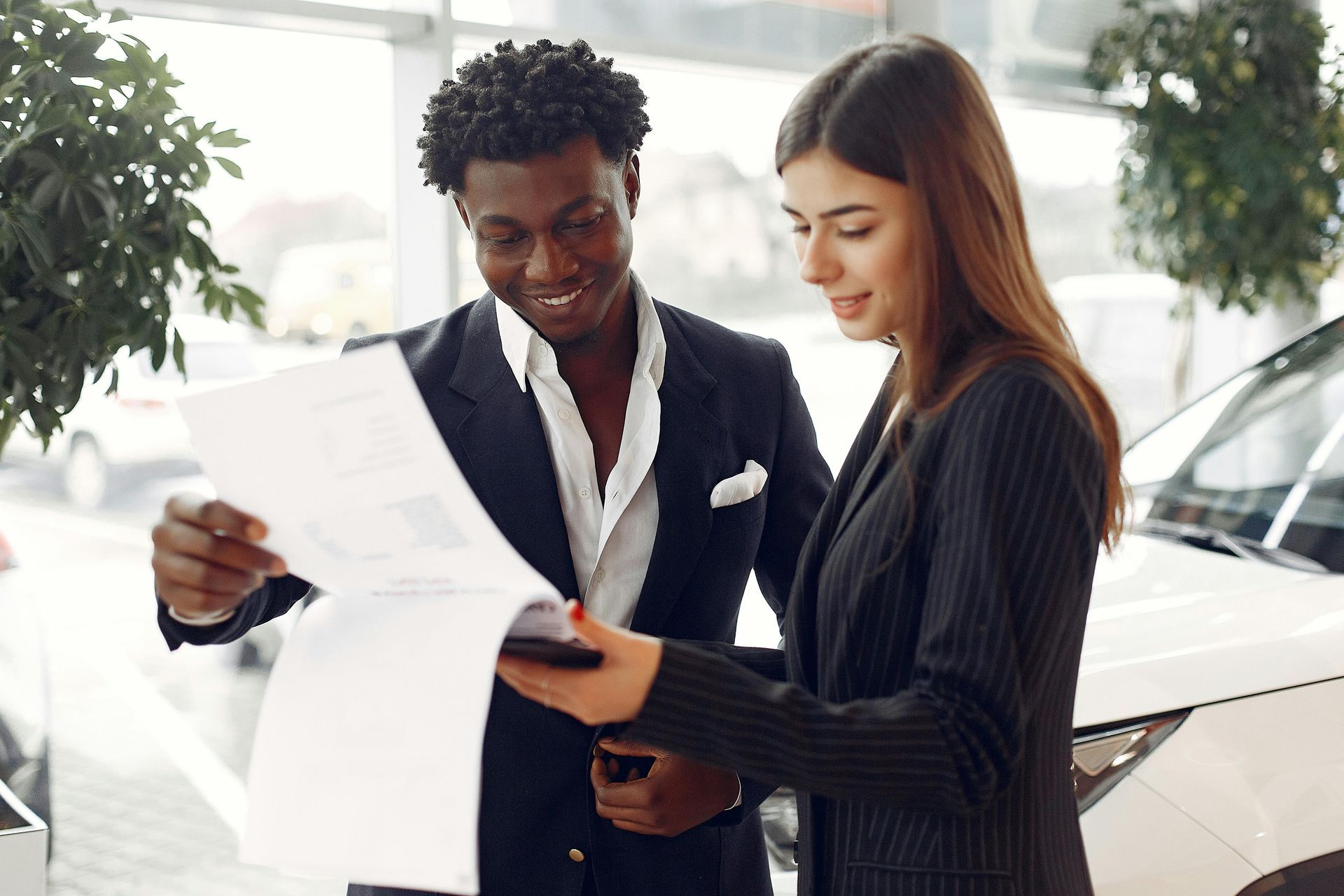 Two colleagues review documents beside a car in a bright showroom.