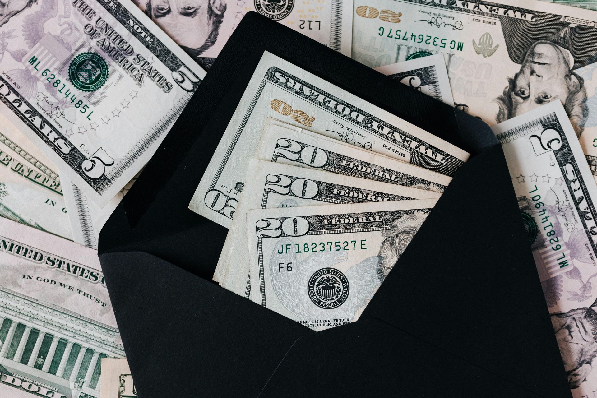 A black envelope sits open, filled with several twenty-dollar bills, surrounded by scattered five and twenty-dollar notes.