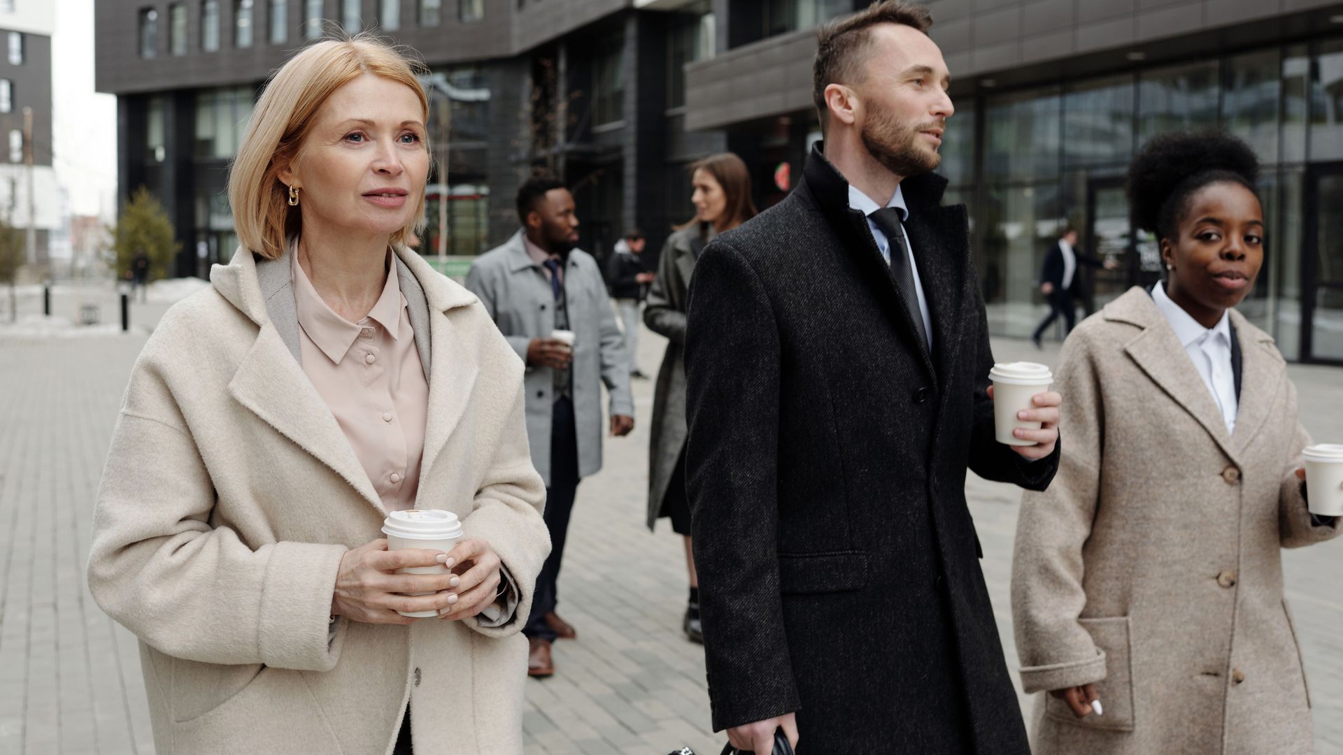 People in business attire walking outside a city building, carrying coffee cups.