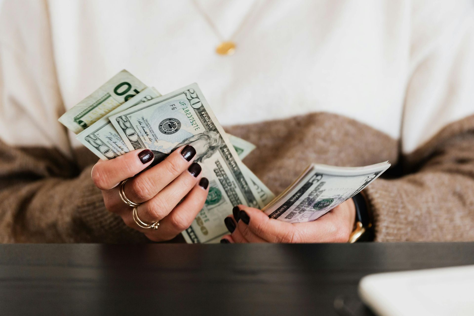 Hands holding and counting US dollar bills. Dark nails, rings, and beige sweater are visible.